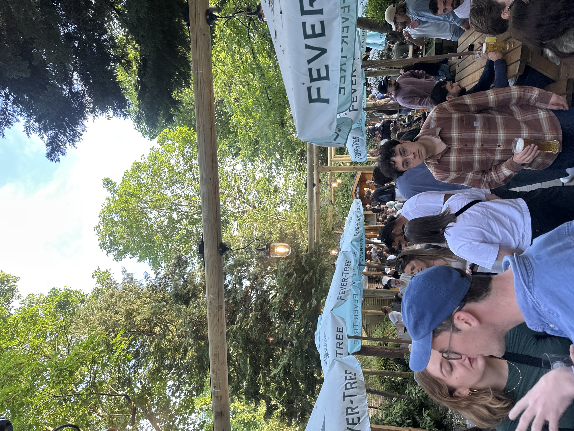 Here is a detailed description of the image:
**Scene**: The image shows a lively outdoor scene, possibly a beer garden or outdoor dining area, likely in London, United Kingdom, given the "Fever-Tree" umbrellas, a brand known for mixers. The area is crowded with people.
**People**: A diverse group of people is present, many seated at wooden tables. They appear to be socializing, eating, and drinking. In the foreground, a couple is kissing. There are people of different ages and appearances.
**Objects**: Tables and benches, presumably for dining, are visible. Above, there are multiple white umbrellas with "Fever-Tree" branding. Light fixtures hang from a wooden structure above the crowd. People are holding glasses, presumably filled with drinks.
**Actions/Interactions**: The primary activity seems to be socializing and enjoying beverages. People are talking, eating, and drinking. The couple in the foreground is kissing.
**Environment**: The scene is outdoors, with trees and foliage visible in the background and above the structure, giving a sense of a garden-like setting. The lighting suggests it might be late afternoon or evening. The wooden structure with light fixtures gives a rustic feel.
