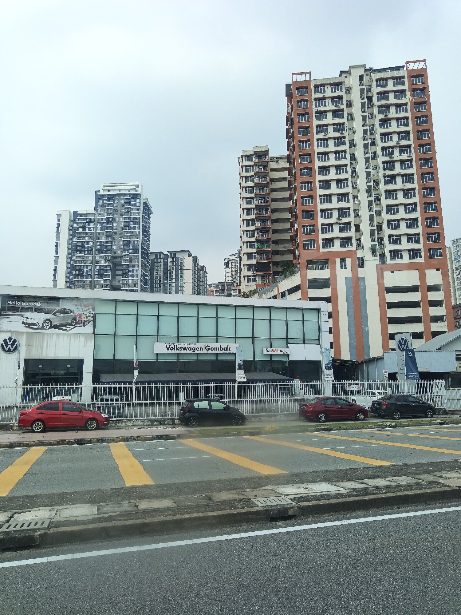 The image shows a car dealership, "Volkswagen Gombak," located in Kuala Lumpur, Malaysia. The dealership features a modern building with large glass windows and prominent Volkswagen branding. Several cars are parked outside, including a red sedan and a black hatchback. In the background, several high-rise apartment buildings are visible against a cloudy sky, characteristic of an urban environment in Kuala Lumpur. The scene depicts a typical street view with a dealership and residential buildings.