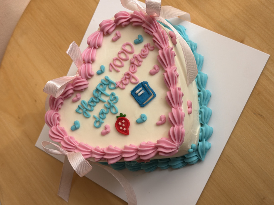 A close-up, high-angle image depicts a heart-shaped cake resting on a white square cake board. The cake is predominantly covered in white frosting. A border of pink piped frosting in a shell or ruffle pattern encircles the top edge of the cake. A second border of light blue piped frosting in a similar pattern encircles the base of the cake. Light pink satin ribbons are positioned around the sides of the cake, with two bows visible on the upper left and right.
The top surface of the cake features various decorations:
- The inscription "Happy days" is piped in light blue frosting.
- The inscription "1000 together" is piped in pink frosting.
- Numerous small, light blue heart-shaped decorations are scattered across the top.
- Numerous small, pink teardrop or elongated heart-shaped decorations are also scattered.
- A single red strawberry decoration with a green stem is positioned on the left side.
- A single blue square-shaped decoration, possibly representing a book or a pause button with two white vertical lines, is located on the right side.
The cake board is situated on a light brown wooden surface, likely a table. No individuals are visible. The image does not contain any discernible elements to identify the specific city location as Tel Aviv-Yafo, Israel.