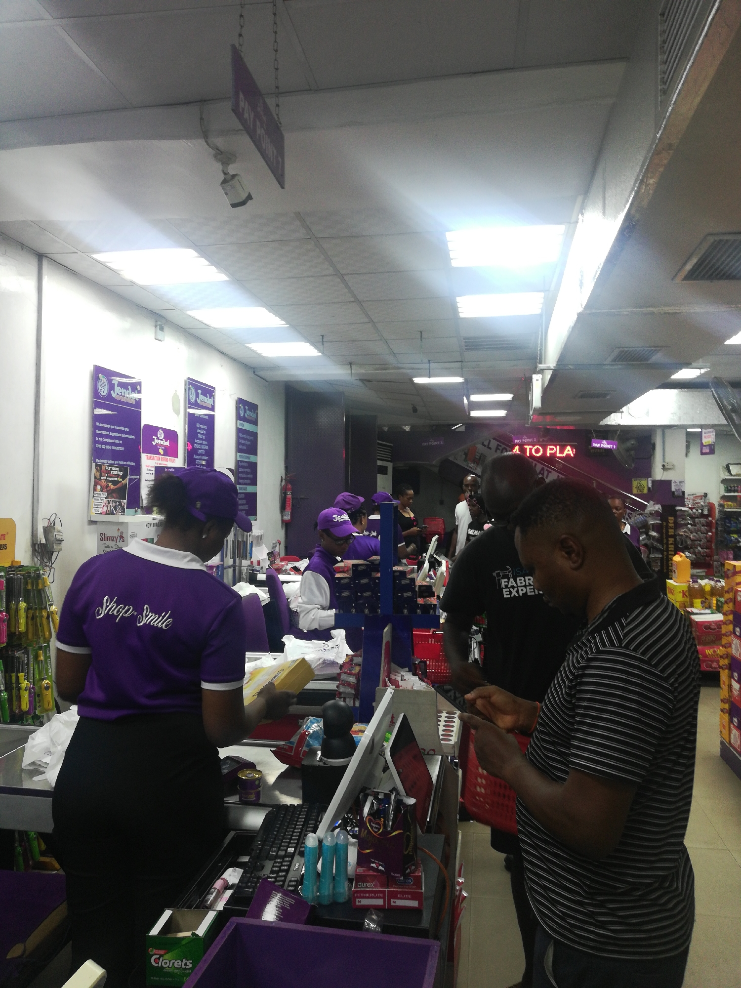 A medium shot captures a supermarket checkout scene in Lagos, Nigeria. The cashier, identifiable by a purple shirt with "Shop Smile" printed on the back and a matching cap, is attending to a customer. Another cashier in a similar uniform is present at the next checkout lane. A man with a red shopping basket waits in line, looking at his phone. Overhead signs read "PAY POINT 3" and "PAY POINT 7" with "ALL FAT TO PLA" above them. The supermarket features overhead fluorescent lighting and shelved goods. Promotional posters for "Jendol" brand are visible on the wall behind the cashiers. "Clorets" gum and "Durex" condoms are on the checkout counter.