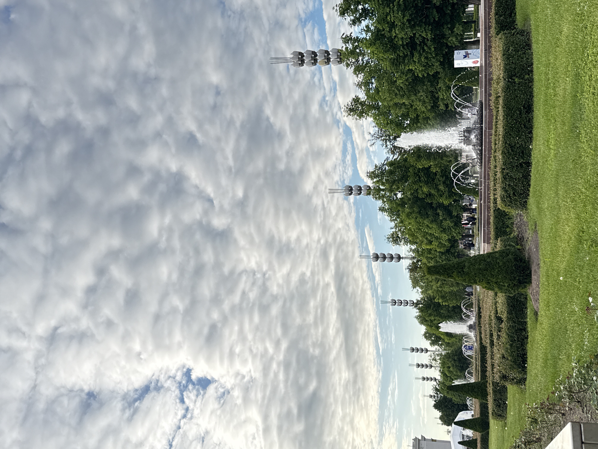 This image captures a wide view of a well-maintained public park or garden, likely in Moscow, Russia, under a partly cloudy sky during the daytime.
The upper portion of the frame is dominated by the sky, displaying a mix of bright white and light grey clouds interspersed with patches of clear blue. Below the sky, a distinctive row of lampposts or decorative light fixtures stretches horizontally across the scene. Each fixture comprises several dark, spherical or bulb-like elements stacked vertically on slender poles, adding a modern aesthetic to the park.
In the mid-ground, a series of fountains are actively spraying water, creating elegant arcs. These fountains are nestled within a long, landscaped strip featuring lush green trees and neatly trimmed hedges. Faint figures of people can be seen in the background near the fountains, suggesting that individuals are enjoying the park. To the right of the fountains, a small white sign is visible, adorned with a blue star and other red/orange symbols, though any specific text is unreadable due to the distance and resolution.
The lower part of the image showcases a meticulously cut green lawn, separated from the fountain area by a darker strip that could be a path or a planting bed. The overall scene exudes a peaceful and orderly atmosphere, characteristic of a well-kept urban green space. The bright lighting indicates good weather.