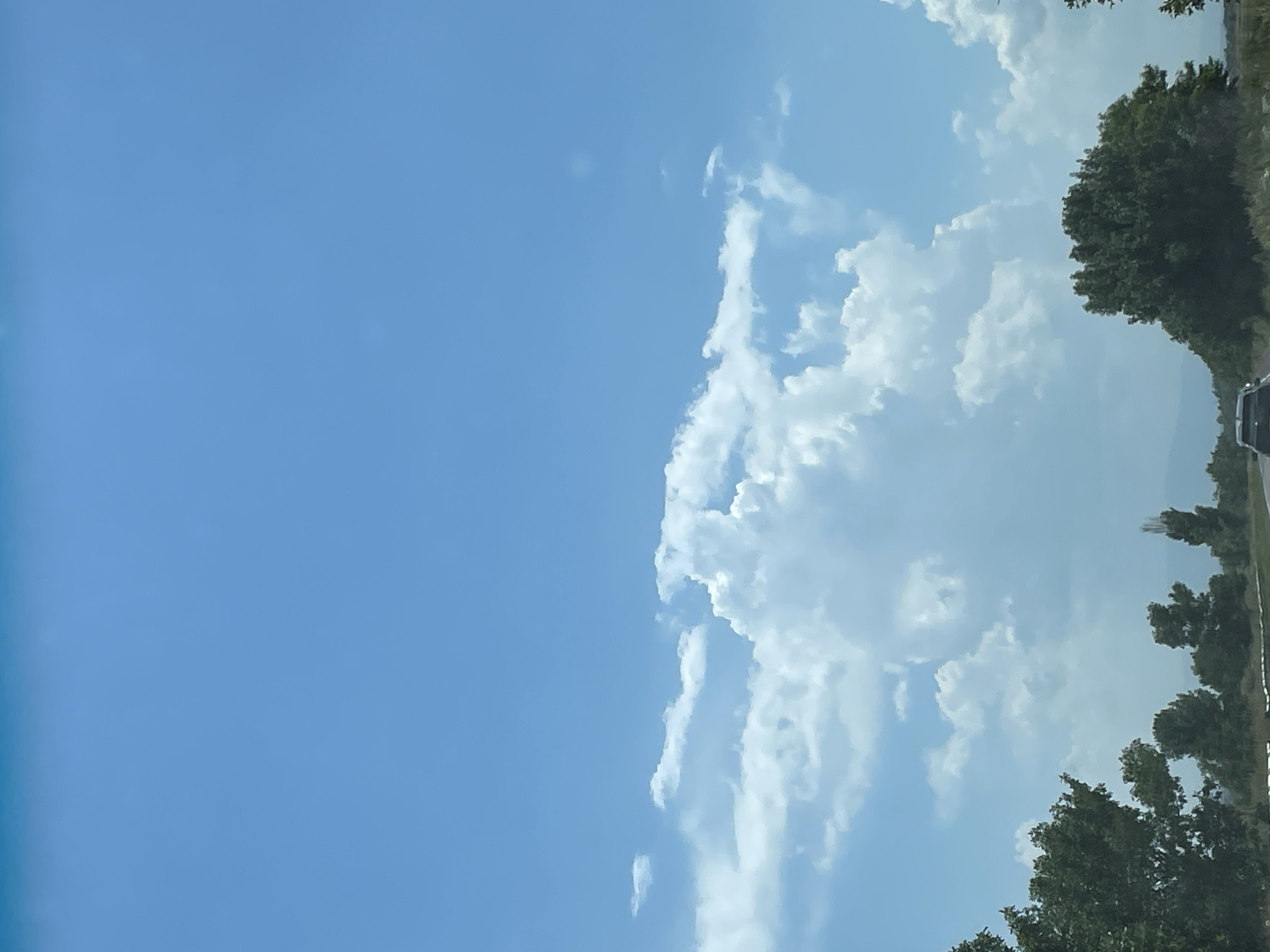 The image depicts a road scene under a bright blue sky with scattered white clouds. In the foreground, a portion of a dark-colored vehicle is visible driving on a road. To the left and right of the road are lush green trees. In the background, a range of mountains is faintly visible. The overall impression is one of a scenic drive in a rural or semi-rural area, likely on a clear day. The specific location in Shirak, Armenia, is suggested by the provided information, though there are no explicit visual cues within the image itself to confirm this.