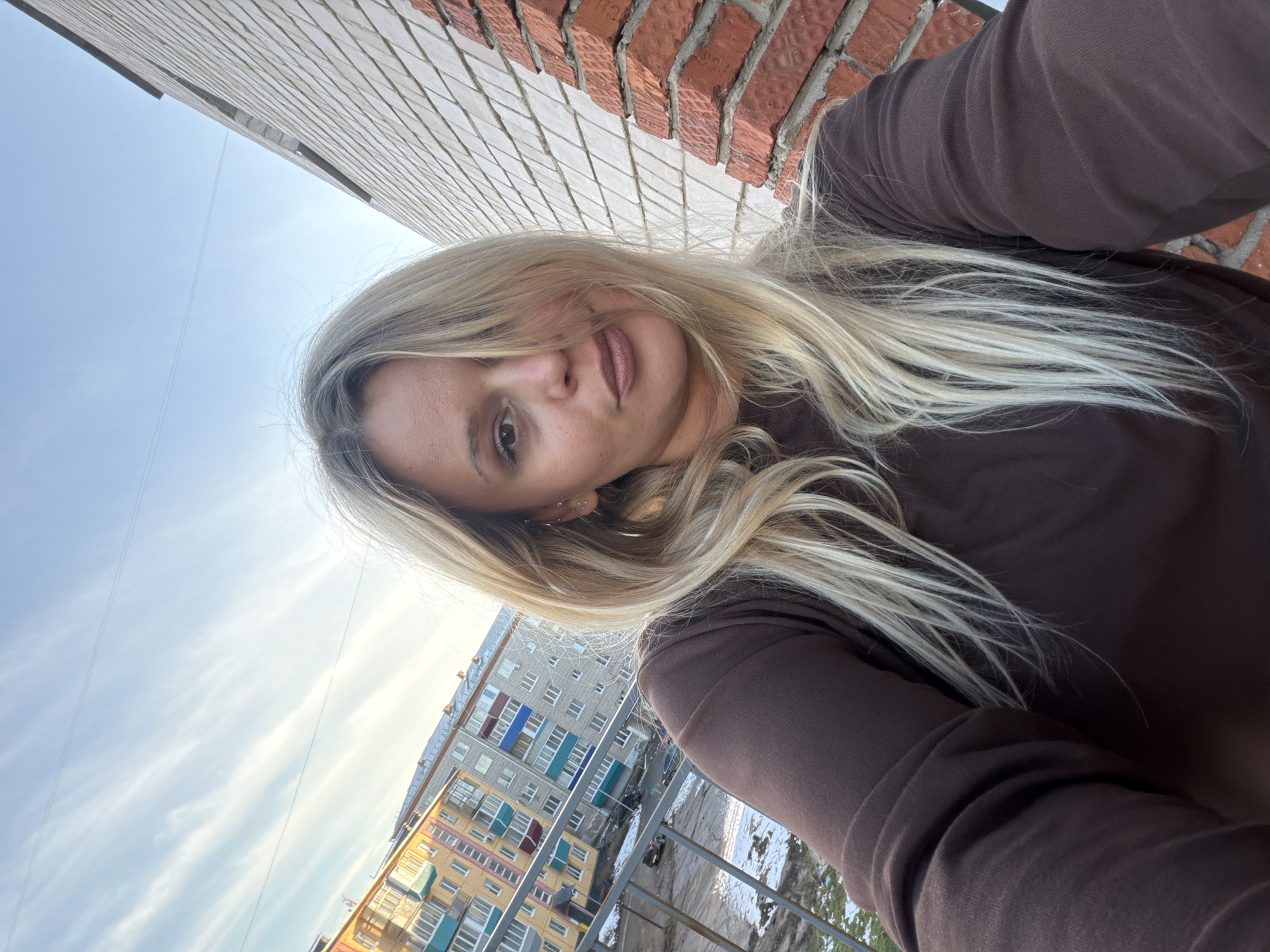 A woman with blonde hair is photographed at eye level, angled with the camera towards her. She is wearing a brown, long-sleeved shirt. Behind her, there is a red brick wall on the right. To the left, there is a white, tiled wall, and a light blue sky with wispy clouds. In the distance, a tall, multi-story building with yellow, blue, and green trim is visible. Below her, a metal railing overlooks a snowy area. Given the provided information, the location is implied to be Sarapul, Russia.