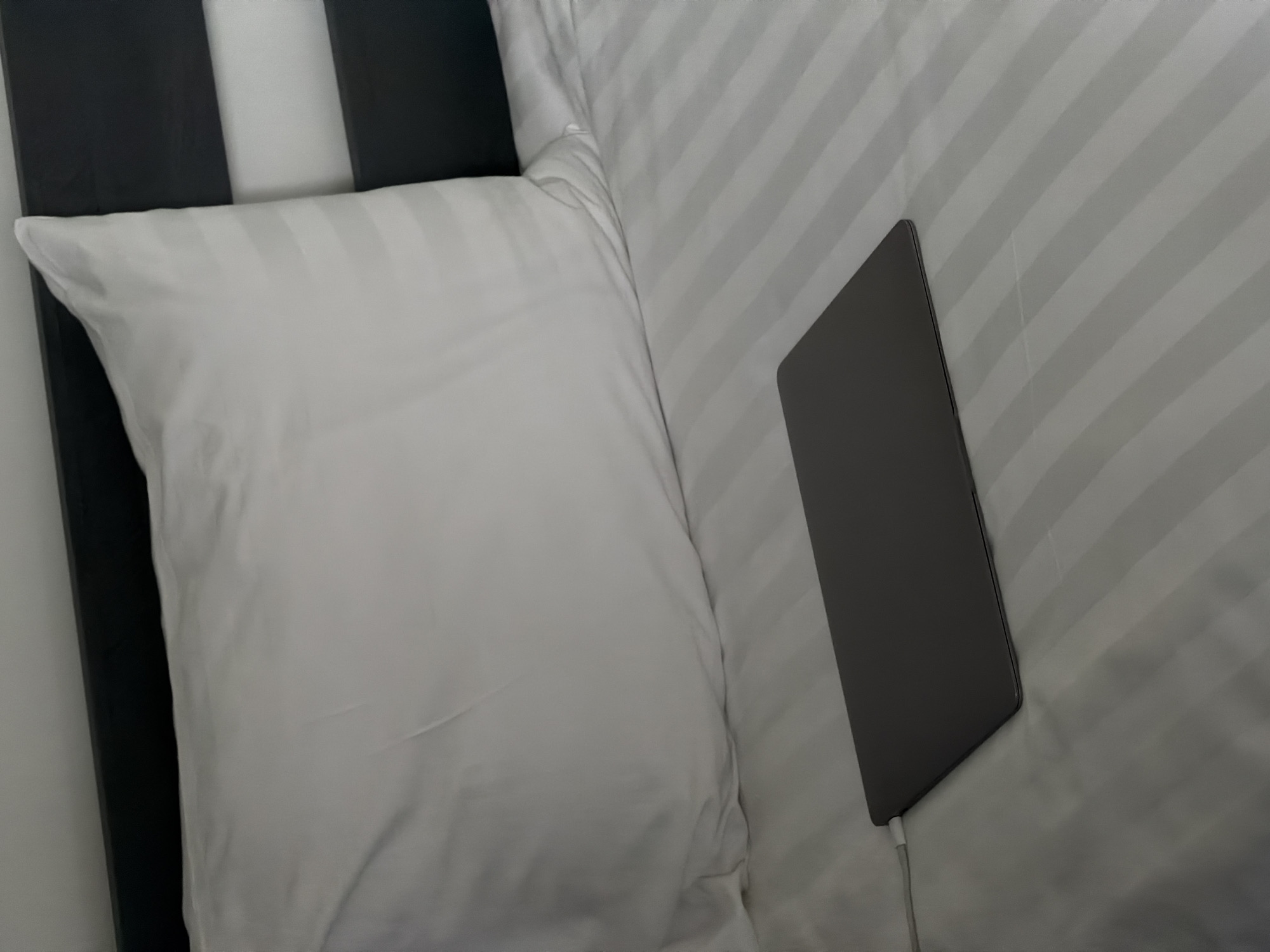 A laptop is plugged in and resting on a striped, white bed. There is a pillow behind it to its left. To the left of the pillow is a black and white headboard. The scene is indoors. There is no indication of this photo being taken in Chkalov, Armenia. No people are present.
