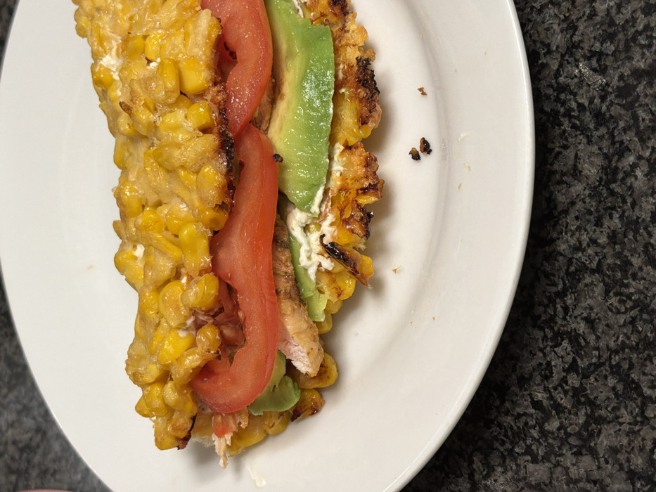 The image displays a close-up view of a single food item, resembling an open-faced sandwich or a type of arepa/taco, resting on a white, oval-shaped ceramic plate. The main structure of the food consists of two flattened, griddled components made primarily from yellow corn kernels, some of which exhibit visible charring. Between these corn layers, the following ingredients are observable: several slices of red tomato, pieces of cooked white meat (consistent with chicken), slices of green avocado, and a white creamy sauce or spread visible between the layers. The white plate is positioned on a dark, mottled countertop, consistent with granite or a similar stone material. Several small, dark food crumbs are present on the white plate near the bottom right edge. No people are visible, and no active actions are depicted. The specific location within the city of Tel-Aviv, Israel, cannot be determined from the visual information present in the image.