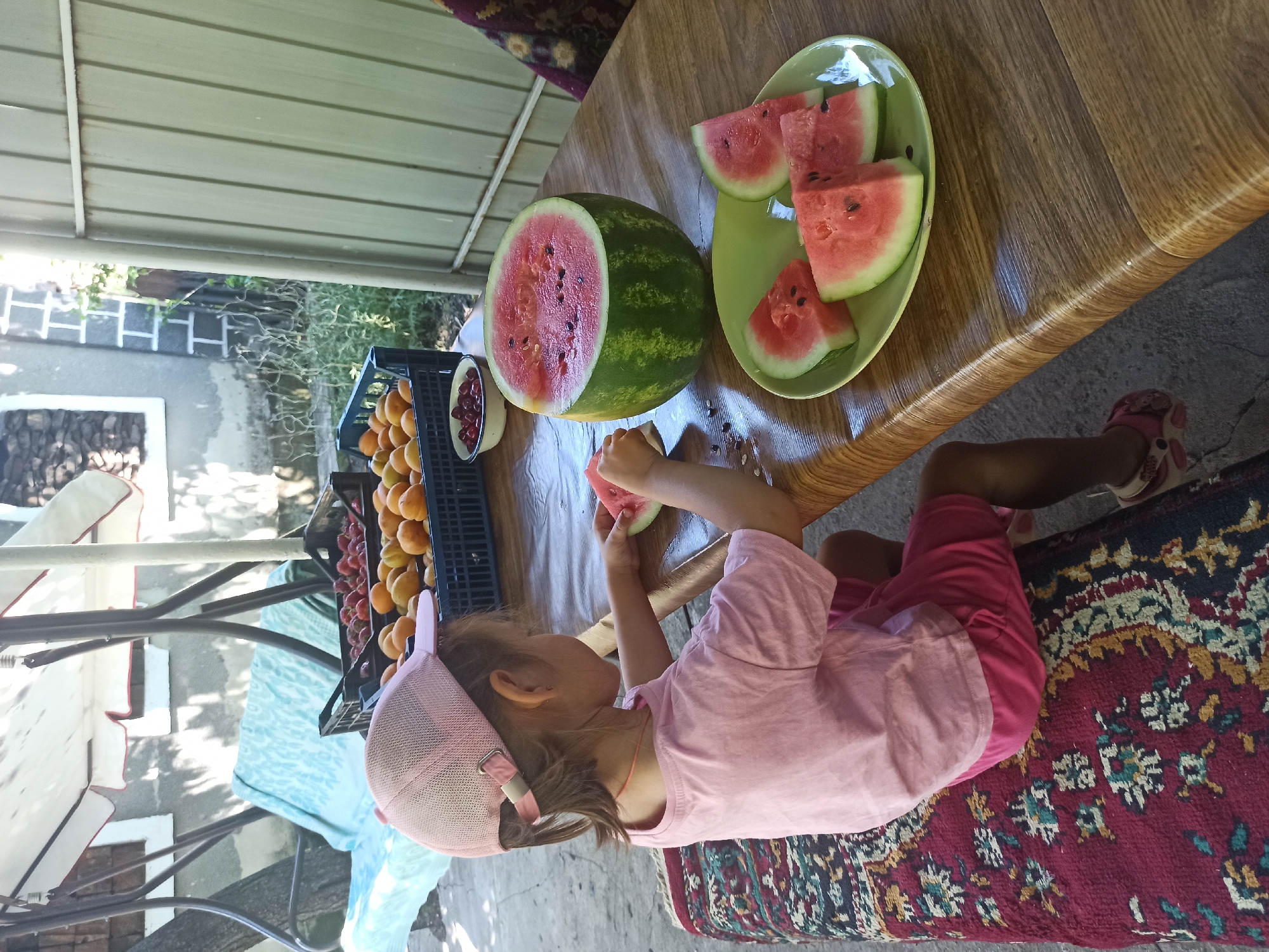 A young girl, wearing a pink cap and t-shirt with pink shorts, sits on a bench covered with a patterned rug. She is seated at a wooden table outdoors, with her back mostly to the camera. In front of her, on the table, is a large, cut watermelon and a plate with several slices of watermelon. A few seeds are scattered on the table. To the left of the table are two black crates filled with fruit, likely apricots and strawberries, and a white bowl with cherries. In the background, there is a swing set with a light-colored canopy and cushions, and a portion of a building with a brick wall. The setting appears to be a yard or patio area. The image was taken in Kryvyi Rih, Ukraine.