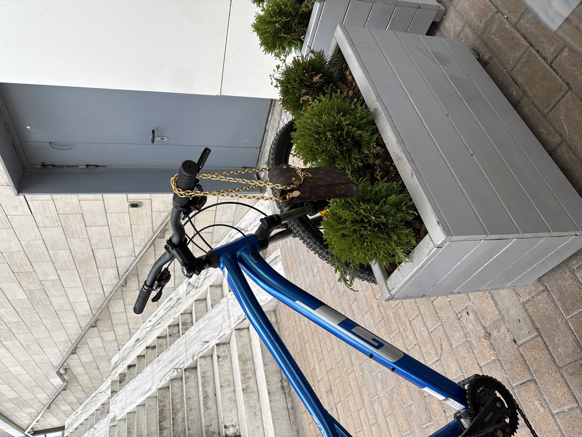 The image depicts an outdoor urban scene in Moscow, Russia, featuring a blue bicycle parked in a well-maintained area. The perspective is slightly elevated, looking downwards at an angle, with the scene appearing bright under daytime lighting.
In the foreground, a glossy bright blue mountain bike is the primary subject. The "GT" brand logo is prominently visible on its frame. The bicycle features black handlebars with black grips and a black front gear mechanism, where the "Shimano" brand name can be read. Hanging from the handlebars is a small brown handbag or pouch with a distinctive monogram pattern, highly resembling Louis Vuitton, attached by a prominent gold-colored chain strap.
To the right of the bicycle, running parallel to it, is a long, light gray wooden planter box filled with lush green, coniferous shrubs, likely thuja. Beyond this planter, the ground is paved with light-colored bricks.
On the left side of the frame, a wide, light-colored tiled staircase descends, suggesting an entrance or transition to a lower level. The tiles are square and uniform. In the upper left background, a modern building facade with white and gray panels is partially visible. The scene is devoid of people, presenting a calm and still environment. The overall setting suggests a clean, contemporary public or semi-public space, such as a plaza or building entrance.