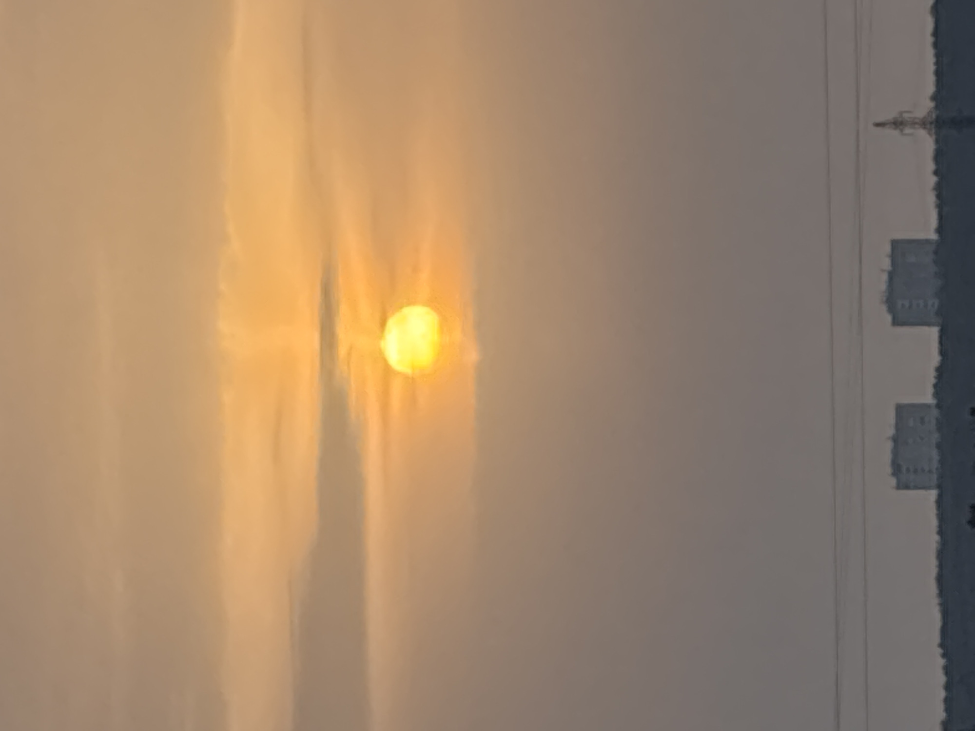 The image presents a panoramic view of a distant urban horizon at either sunrise or sunset, set in Chaikovsky, Russia. The upper two-thirds of the frame are dominated by a sky displaying a gradient of warm yellowish-orange hues transitioning to a muted orange-grey. The sun, appearing as a distinct, intensely yellow spherical orb, is positioned in the upper-left quadrant, partially obscured by diffuse, light-colored clouds, which are illuminated by its glow. Below the sky, a dark, indistinct treeline forms a horizontal band across the lower portion of the image. Above this treeline, two light-colored, multi-story rectangular buildings, consistent with residential structures, are visible in the mid-ground, silhouetted against the lighter sky. To the far right, an electrical transmission pylon is discernible as a dark silhouette. Two thin, dark horizontal lines, identified as overhead power lines, traverse the lower-middle section of the sky, running parallel above the treeline and distant buildings.