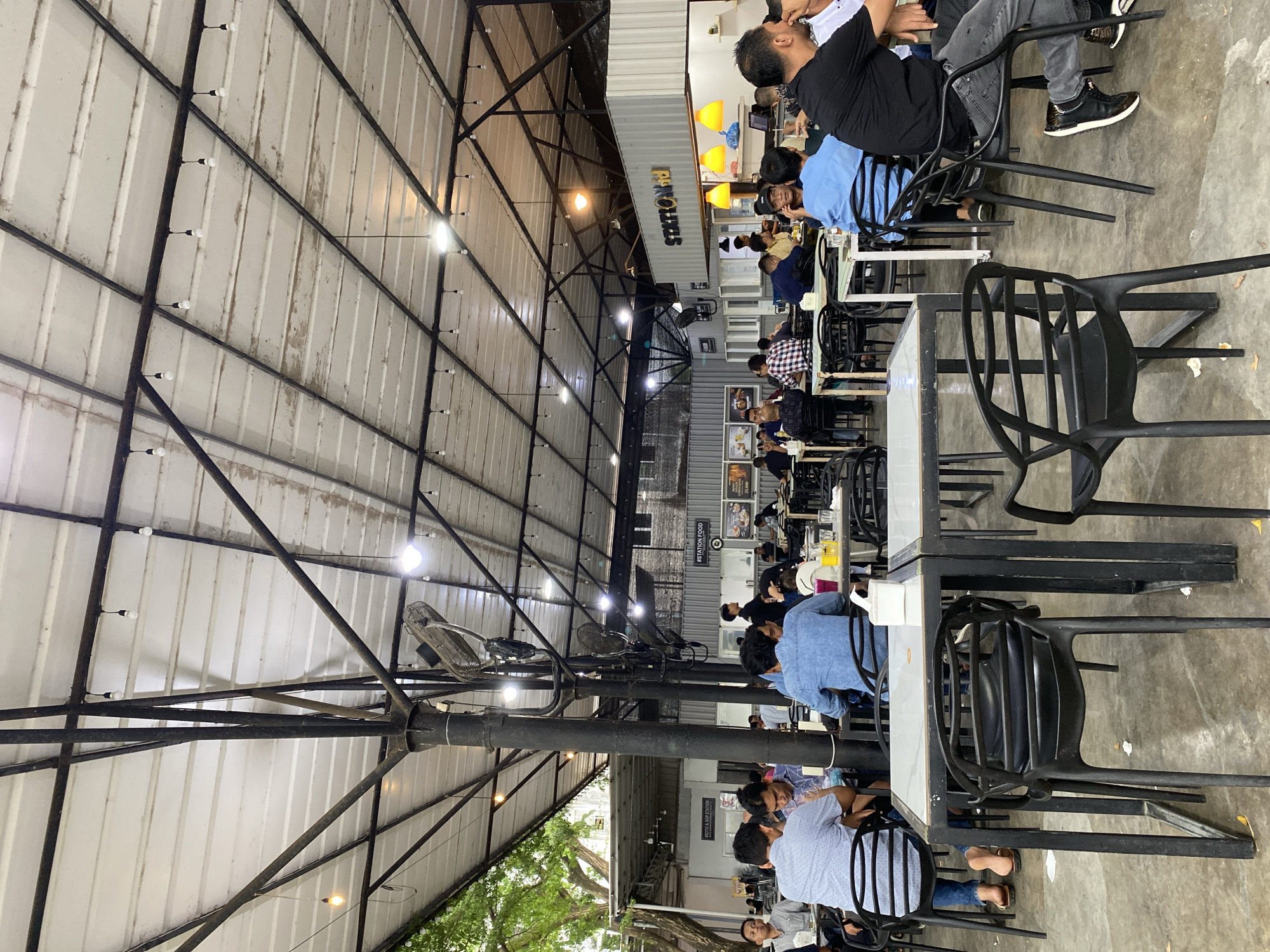 This media file captures a bustling, casual dining area, likely a food court or open-air restaurant in Lhokseumawe, Indonesia. Numerous people, predominantly men, are seated at rectangular tables with black frames and light-colored tops, engaging in conversation and dining. They are dressed in casual attire, including t-shirts, jeans, and denim shirts.
The setting is characterized by an industrial-chic aesthetic, featuring a high, vaulted ceiling constructed from corrugated metal supported by exposed black metal trusses and large vertical beams. Several small, bare lightbulbs are strung along the ceiling beams, complementing larger ceiling lights and a few yellow-orange pendant lamps visible near a serving counter. Industrial-style ceiling fans are also mounted to provide ventilation.
In the background, a serving counter or kitchen area is visible, prominently displaying the text "STATION FOOD." Below it, a vertical sign reads "#STATION FOOD" and a smaller sign states "MISTER & MR. STATION." Various posters and menus are affixed to the walls, though their specific text is not clearly legible.
The presence of artificial lighting suggests the time of day is likely evening or night. The open structure of the dining area, with trees and foliage visible outside, implies a comfortable climate. People appear relaxed and sociable, enjoying their meals and company in this lively establishment. The flooring is concrete.