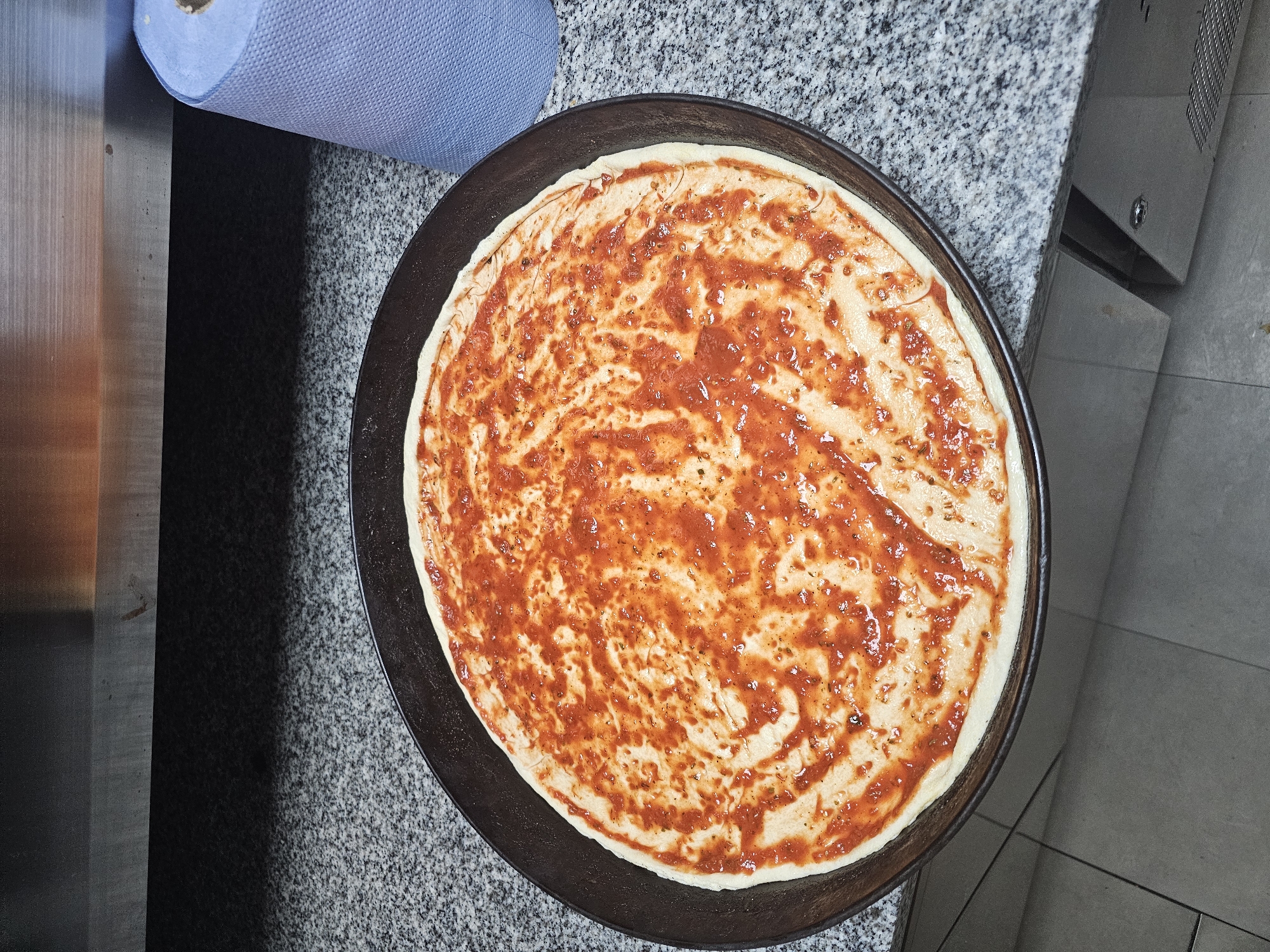 The image shows a pizza crust on a dark metal pizza pan covered with red sauce. The pan rests on a speckled gray countertop next to a roll of blue paper towels and a metal cabinet. The location is unknown, but it is suspected to be in Holywell, United Kingdom. No people are visible.