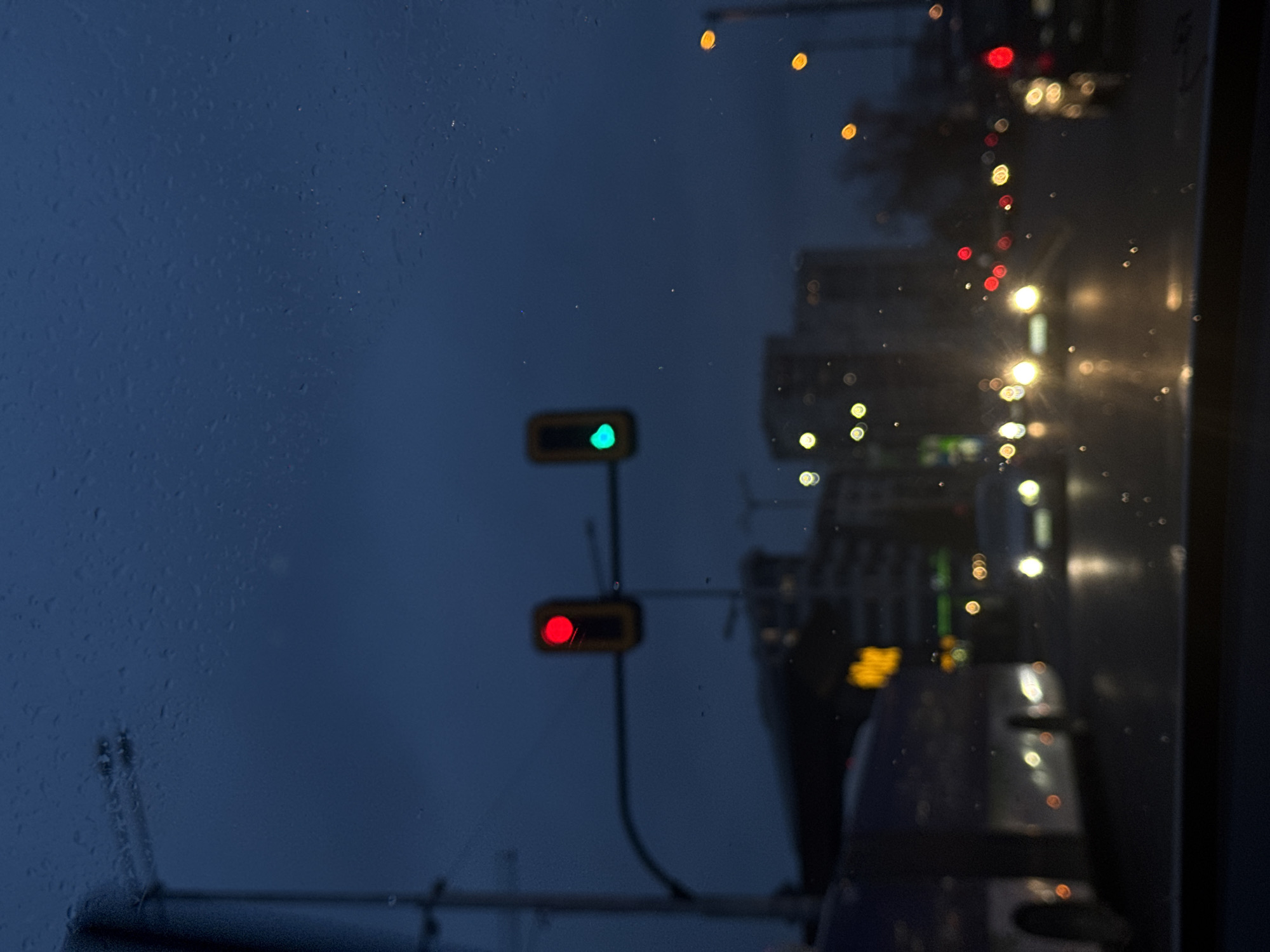 The image appears to be taken from inside a vehicle, looking out at a rainy cityscape at night. On the left, raindrops are visible on the glass. A traffic light is visible in the mid-ground; the top light is green, and the bottom light is red. Beyond the traffic light, there are blurred lights indicating other vehicles and buildings, suggesting a city environment. The lights include street lights, headlights, and possibly building lights. The general lighting and blurred details suggest it is dark. There is no indication of location such as Sofia, Bulgaria from the information presented in the picture. No people are visible.