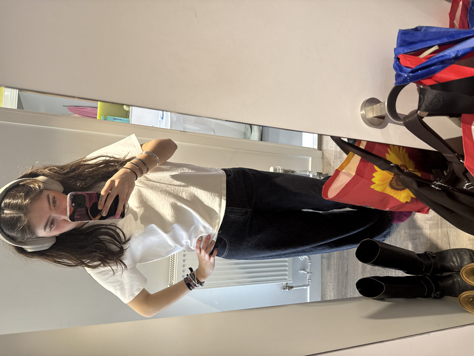 A young female is depicted in a mirror selfie, standing indoors in what appears to be a residential setting in Hull, United Kingdom. She has dark, wavy hair, parted centrally, and wears light-colored over-ear headphones. Her attire includes a white short-sleeved t-shirt and dark, wide-leg trousers. Multiple bracelets are visible on both wrists: several multi-colored bands on the left, and several thin metallic bracelets on the right. She holds a dark-cased smartphone in her right hand to capture the reflection, with her left hand placed on her hip. The floor is light-toned, possibly laminate. To her left, a white panel radiator with exposed pipework is mounted on a white wall. At the base of the mirror, on the right side from the viewer's perspective, a red reusable shopping bag featuring a yellow sunflower design, a black handbag, and other fabric bags (including red and blue) are situated on the floor. To her left, a pair of black mid-calf boots with buckle details are also on the floor. The background reflection reveals a slightly ajar white door with a silver lever handle, opening to an adjacent room, where a white sink and colored textiles (possibly towels) are partially visible, suggesting a bathroom or utility area. No specific landmarks within Hull are discernible from the image itself.