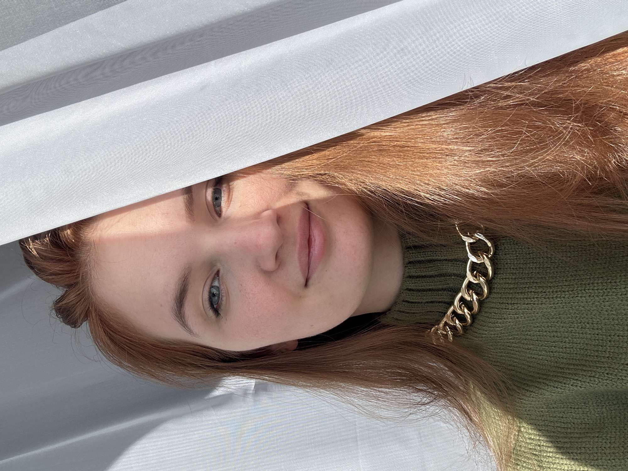 A fair-skinned woman with light auburn hair is depicted in this portrait. The woman is wearing a green turtleneck sweater and a gold chain necklace. Her expression is a soft smile and her blue eyes are visible. Part of her face is obscured by a white, sheer fabric, possibly a curtain or blind. The lighting suggests an indoor setting, possibly near a window. No information indicates the location is in the city of Chaikovsky, Russia.