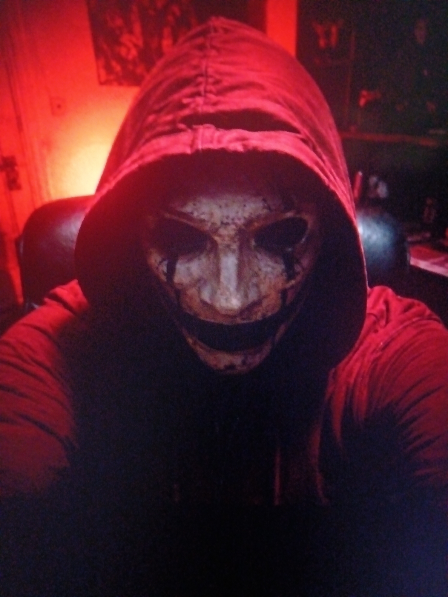 An individual wearing a red hooded sweatshirt and a white mask with dark eye holes and a jagged mouth is positioned in the foreground of the image. The mask has a distressed, textured appearance, with dark smudges around the eye and mouth areas. The individual's face is not visible. The background is dimly lit with a prominent red hue, possibly from ambient lighting. To the left of the individual, a portion of what appears to be a dark chair is visible. In the upper left corner of the background, a framed image or poster with abstract shapes can be discerned. The overall lighting conditions create a dark and somewhat ominous atmosphere. There is no discernible connection to the city of Alaverdi, Armenia, within the visual information provided.