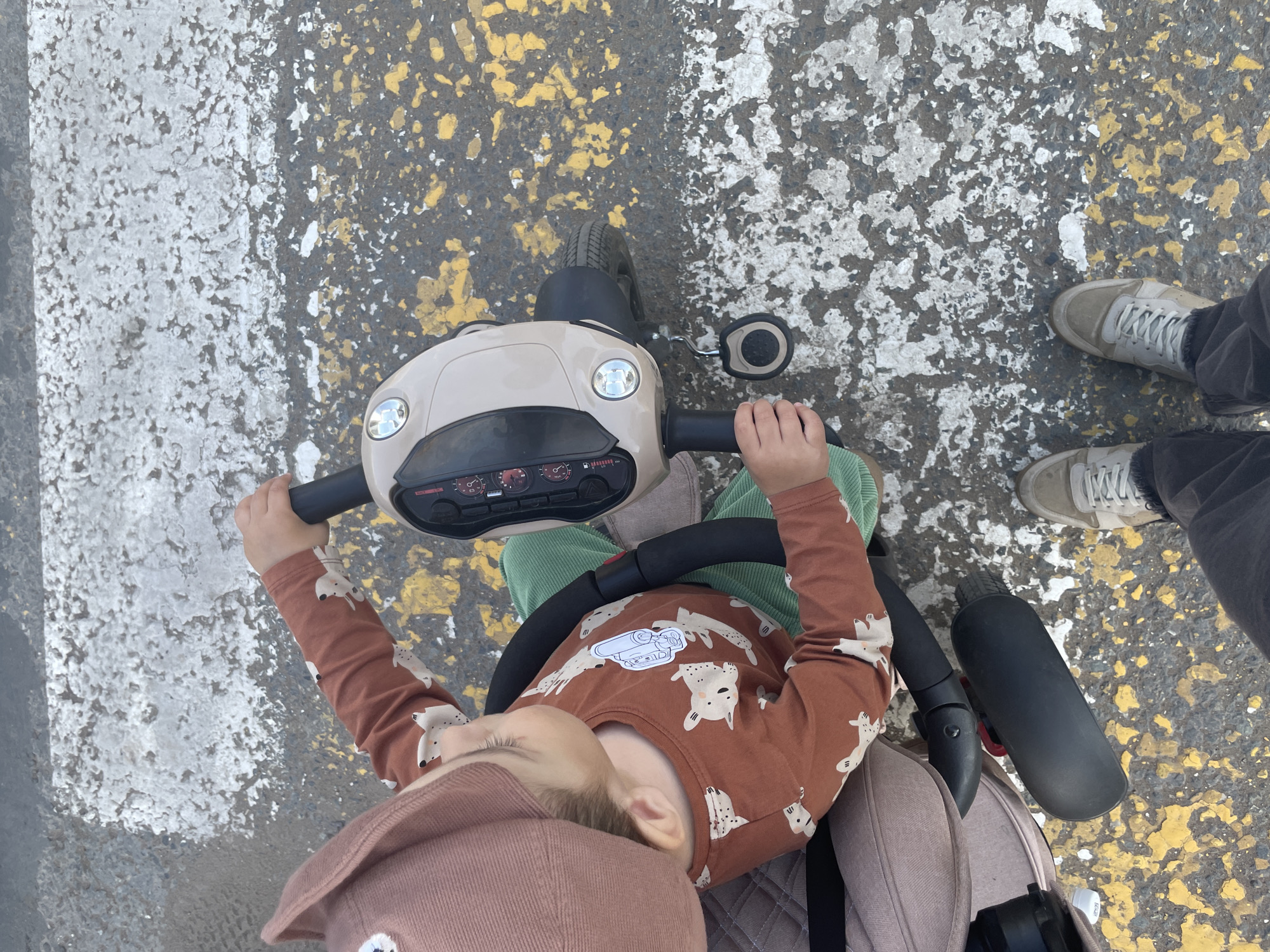An overhead, high-angle shot captures a young child sitting in a beige and black stroller, positioned on a crosswalk with worn white and yellow markings on asphalt. The child, wearing a brown long-sleeved shirt with a white cartoon animal print and a matching pink hat, is holding onto the stroller's black handlebar. The child's left hand is visible, resting on the handlebar. A portion of an adult's legs, clad in dark pants and light-colored sneakers, is visible at the bottom of the frame, suggesting the adult is standing beside the stroller. The stroller itself has a beige canopy and a black seat with a grey quilted fabric lining. The background consists of the asphalt crosswalk, showing signs of wear and tear with faded paint. The overall scene appears to be an outdoor urban environment, likely in Izhevsk, Russia, given the context of the prompt, with the child either about to cross or having just crossed the street.