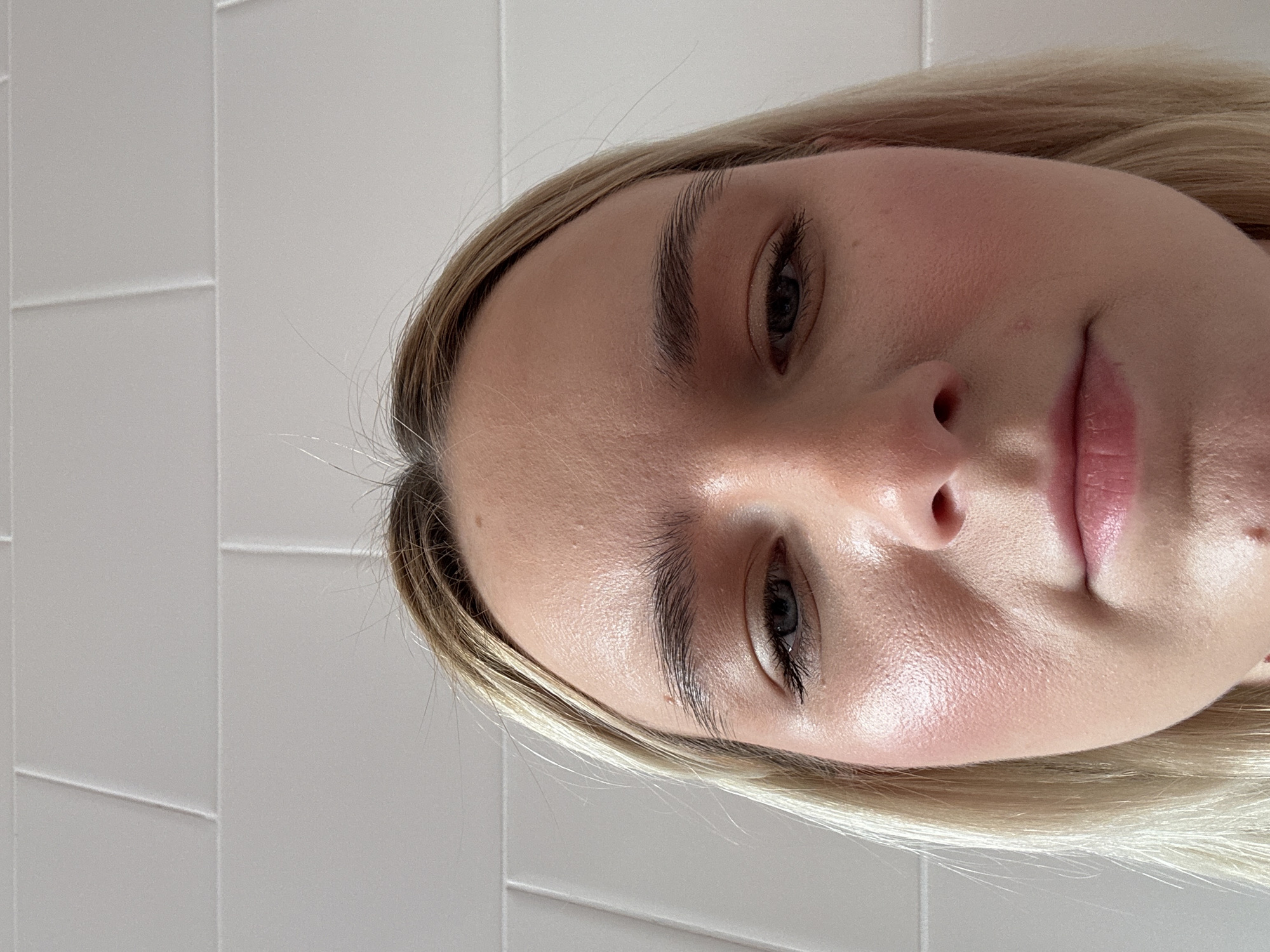 A close-up, slightly angled headshot photograph shows a fair-skinned, young woman with blonde hair. The subject's face appears shiny and moisturized. She is wearing minimal makeup. The background consists of a white tiled wall. The image does not contain information related to Saint Petersburg, Russia.
