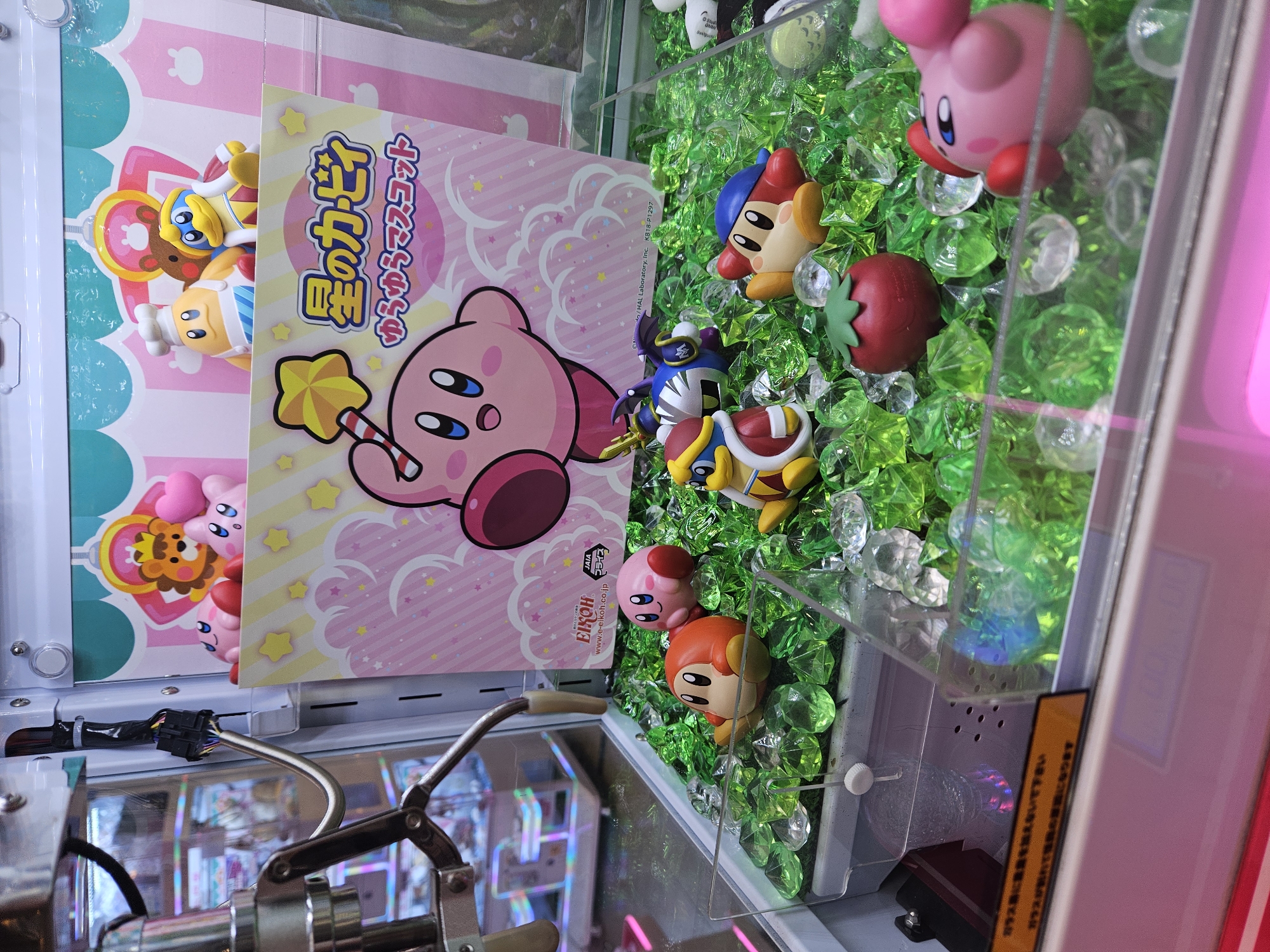 The image shows a close-up of a crane game, filled with Kirby figurines and a promotional poster. The crane's grabbing mechanism is visible in the bottom left, above a mirrored surface. The figurines, arranged on a bed of artificial green grass and clear plastic gems, include multiple versions of Kirby in various poses and costumes. A cardboard advertisement poster features a pink Kirby holding a star wand, with Japanese text indicating "Hoshi no Kirby" (Kirby of the Stars). There are several other smaller promotional posters and figurines in the background, which include more figures with hearts. The general location of the scene is a gaming establishment or arcade, most likely in Japan, considering the Japanese text.
