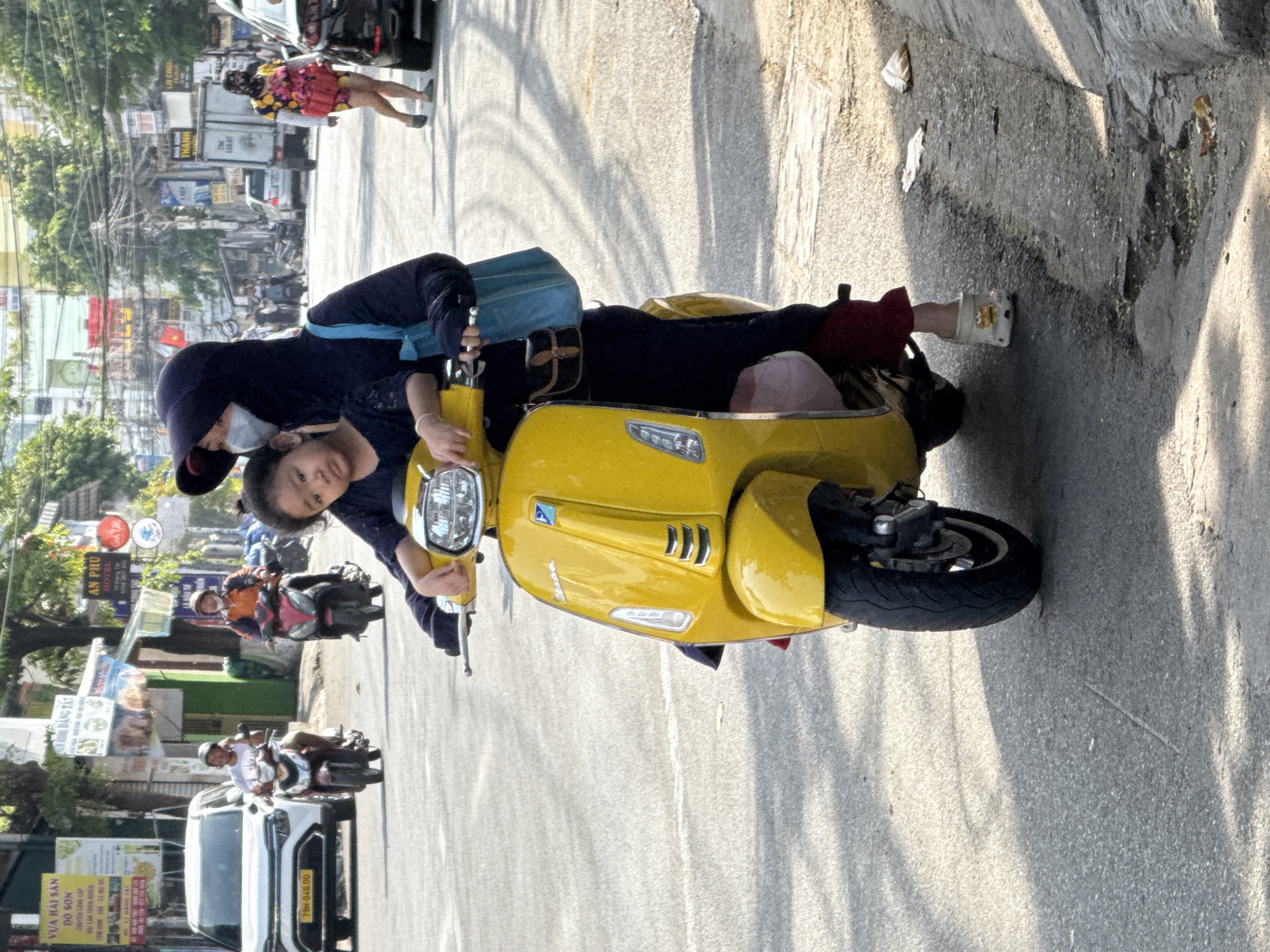 A woman wearing a dark hooded jacket and a face mask is riding a yellow scooter. She is holding the handlebars and a young girl is sitting in front of her, also wearing a dark top. The girl is looking towards the camera. The scooter is on an asphalt road in what appears to be a city. In the background, there are other vehicles, buildings, trees, and power lines. The lighting suggests it is daytime, with shadows visible on the road. The scene is consistent with a street in Bac Nha Trang Ward, Vietnam, given the presence of scooters, local signage and building styles often found in such locations.