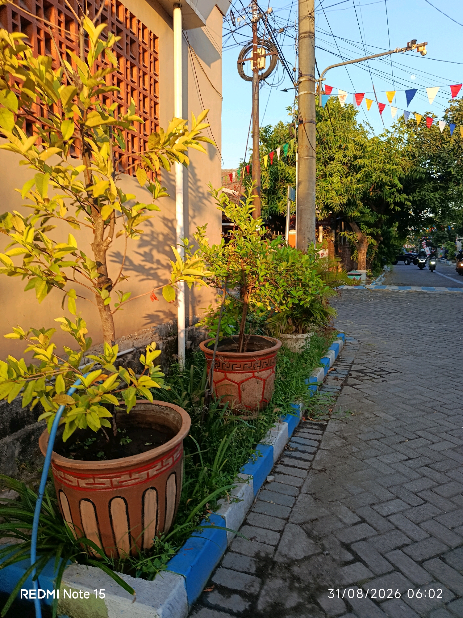 The image depicts a street scene in Keboansikep, Indonesia, at dawn. The foreground features two potted plants placed along a paved walkway with a blue and white border. The plant on the left is taller with yellowing leaves, while the one on the right is shorter and bushier with green foliage. A building with a decorative window grate stands to the left, casting shadows on its wall. In the background, a street lined with trees and electrical poles stretches into the distance. Colorful triangular flags are strung between the poles, suggesting a festive occasion. The paved road shows signs of recent wetness, with reflections visible. Two motorcycles are visible on the road, indicating ongoing traffic. The sky is clear and blue, illuminated by the rising sun. The timestamp on the image indicates the date 31/08/2026 and the time 06:02. The text "REDMI Note 15" is also present at the bottom left.