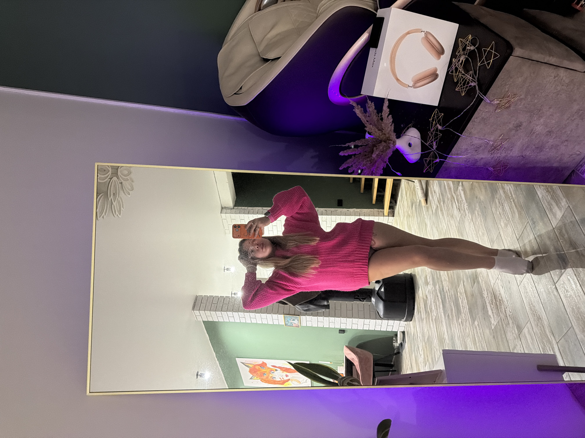 A reflection is captured in a tall, ornate mirror. The reflected image shows a woman with long blonde hair, wearing a bright pink sweater, and holding a smartphone up to take a selfie. The reflection reveals a room with a predominantly green wall, accented with white brick and decorative paintings. The surrounding environment of the room includes various pieces of furniture and decor, such as a grey chair, a table with headphones in a box, and star-shaped string lights. It is unknown if the location is in Большая Шильна, Russia.