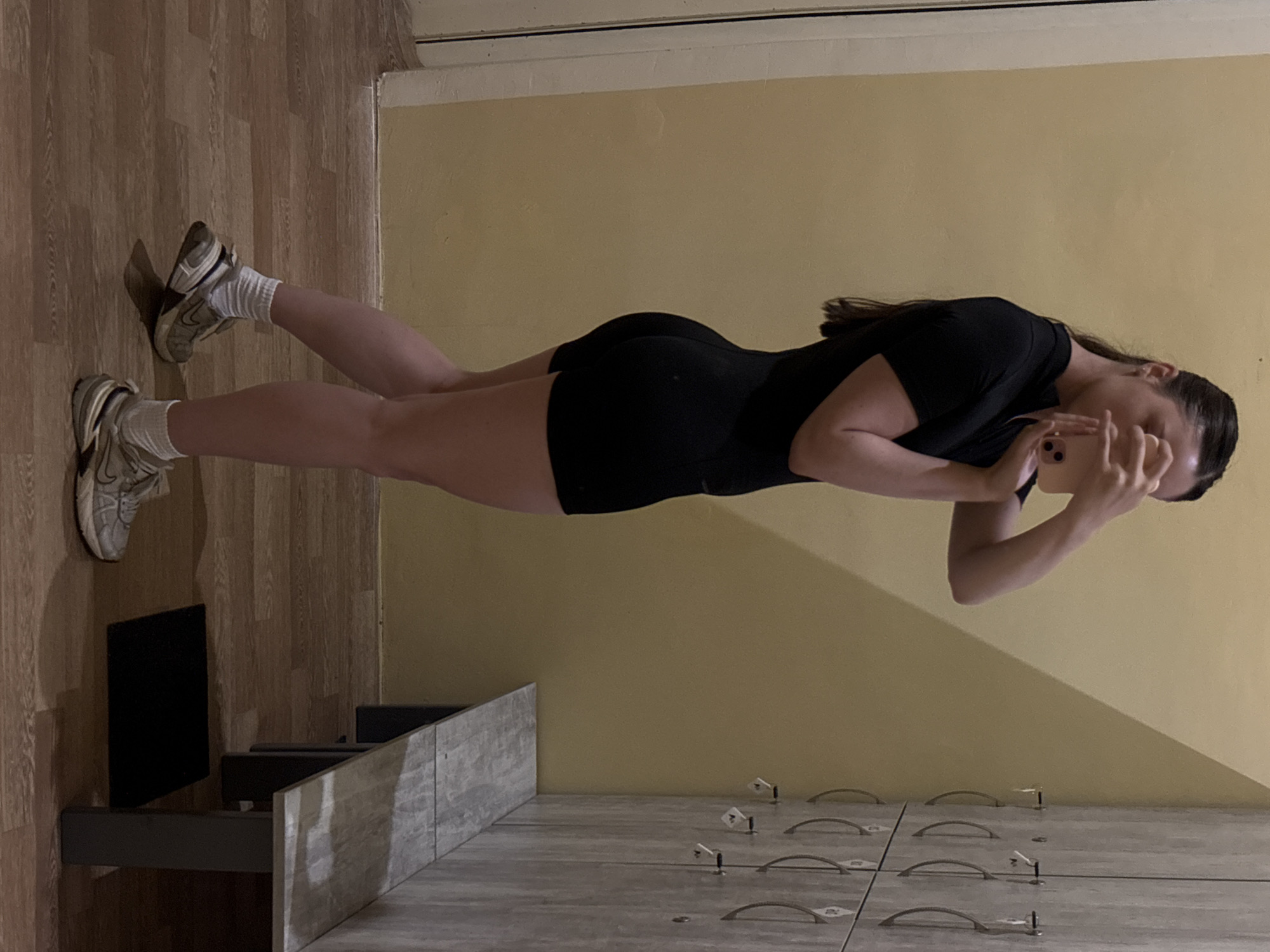 In a locker room, a young woman with dark hair pulled back in a ponytail is captured mid-stride, holding a phone to her face and presumably taking a selfie. She is wearing a black athletic outfit consisting of a short-sleeved top and form-fitting shorts. Her posture is dynamic, with her left leg extended slightly backward, showcasing her athletic physique. She has white socks and athletic shoes on her feet. The background features a wall with a distinctive yellow hue and a row of light-colored lockers, each with a metal handle. A wooden bench with a stone-like pattern is positioned in front of the lockers, and a dark mat is visible on the floor beneath it. The flooring is made of wooden planks.
