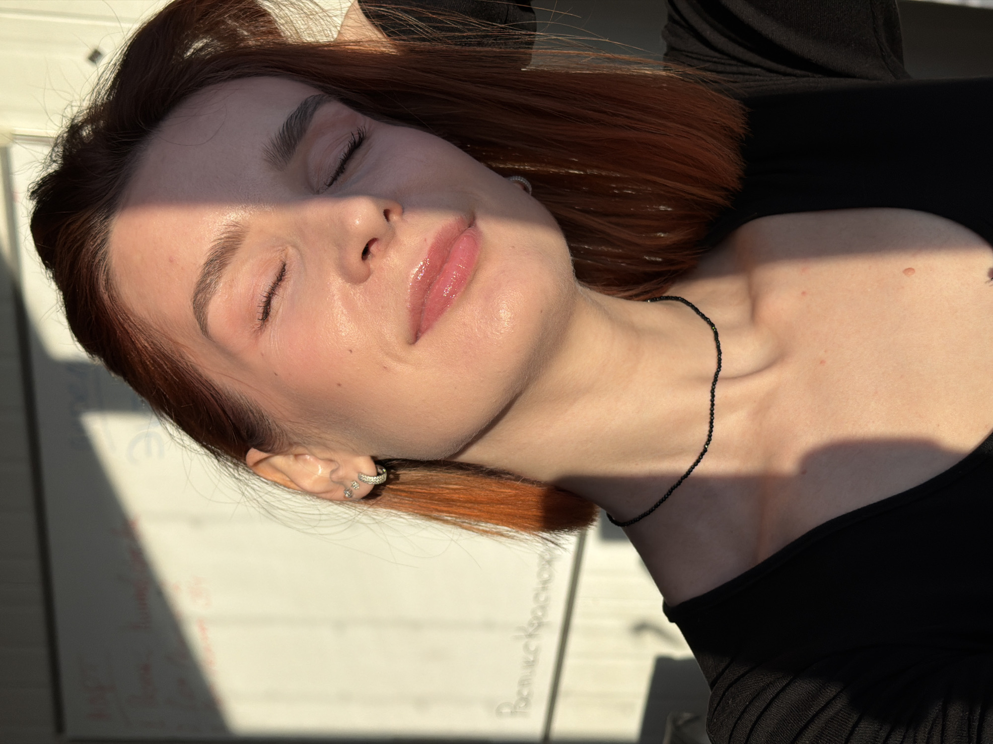 A woman with red hair and closed eyes is photographed in bright sunlight. She wears a black top and a beaded necklace. She has multiple earrings in her left ear. The location is possibly Chaikovsky, Russia, based on the Cyrillic script visible on a surface next to her.