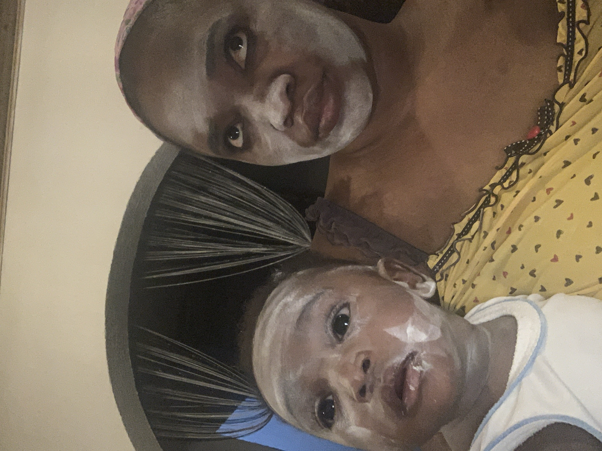 The image shows two individuals with a white substance applied to their faces, likely a facial treatment. An adult female with a pink head wrap and a yellow heart-patterned top is positioned above a young child wearing a white shirt with blue trim. The child has a ponytail. The background includes a neutral-colored wall and a dark-colored circular object with radiating lines. There is no information in the image to indicate the location in the city of Barakin Akawo, Nigeria.