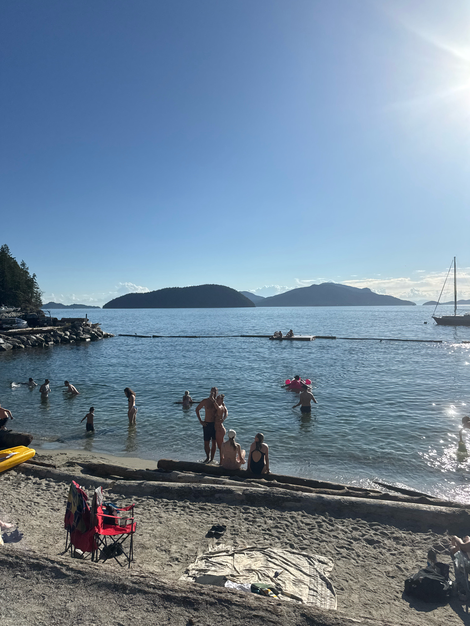 The image depicts a sunny day at a beach in Lions Bay, Canada. The scene is populated with numerous people enjoying the water and the shore. In the water, individuals are swimming, wading, and some are on inflatable toys. A group of people are gathered on a floating platform further out in the water. On the beach, there are beach chairs, towels, and logs. A yellow kayak is visible on the sand. A sailboat is anchored in the distance. The background features a forested coastline and mountainous islands under a clear blue sky with the sun shining brightly.