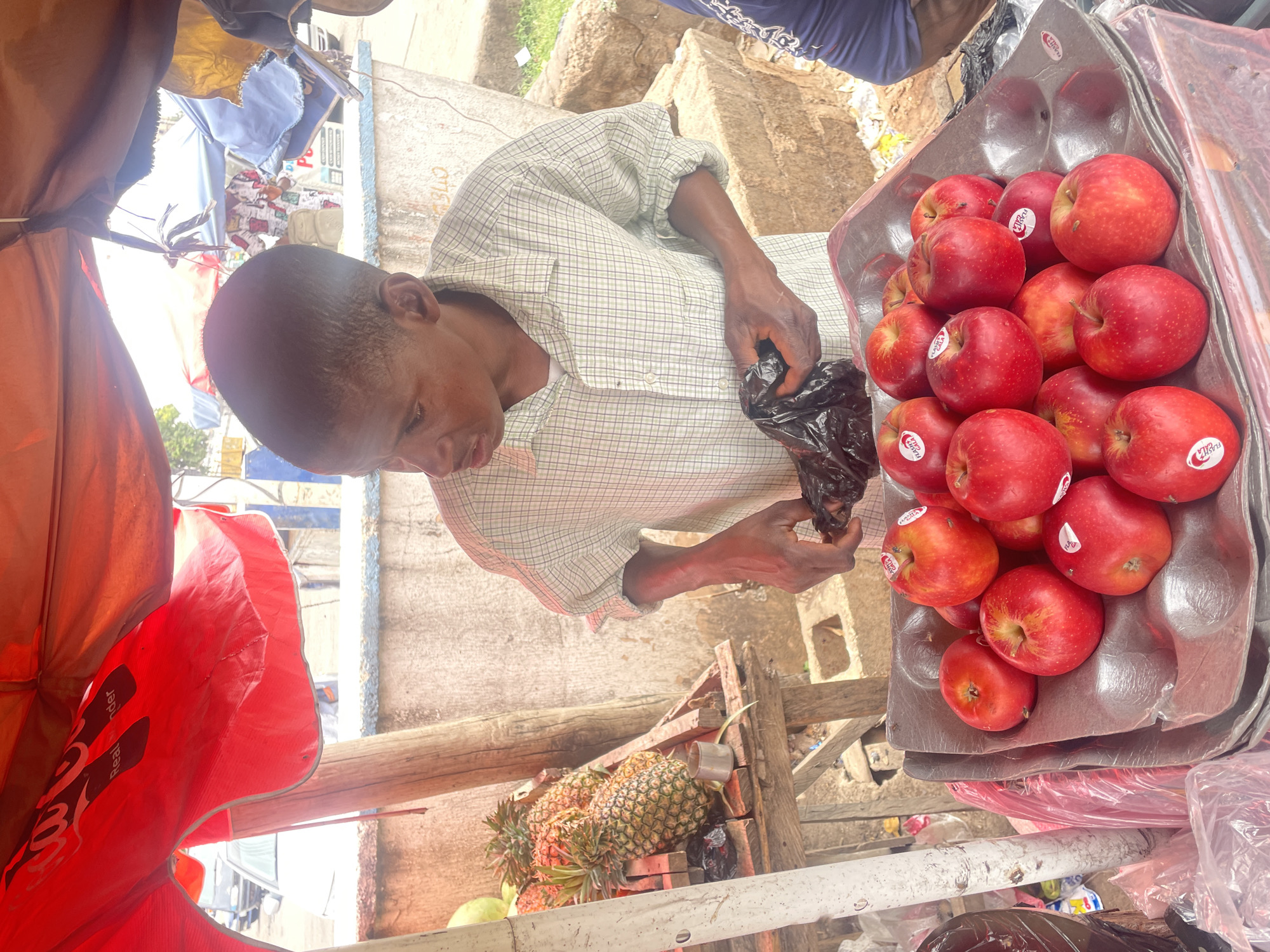 The image depicts a man at a fruit stand in Jos, Nigeria. He is inspecting something in a black plastic bag. Next to him is a tray filled with red apples, each bearing a sticker. Underneath the apples are pineapples. The man is wearing a light-colored, checkered shirt. The scene is set in an outdoor market area with elements of a vendor's stand, including a red and white canopy. The surroundings appear to be a street or roadside market setting.