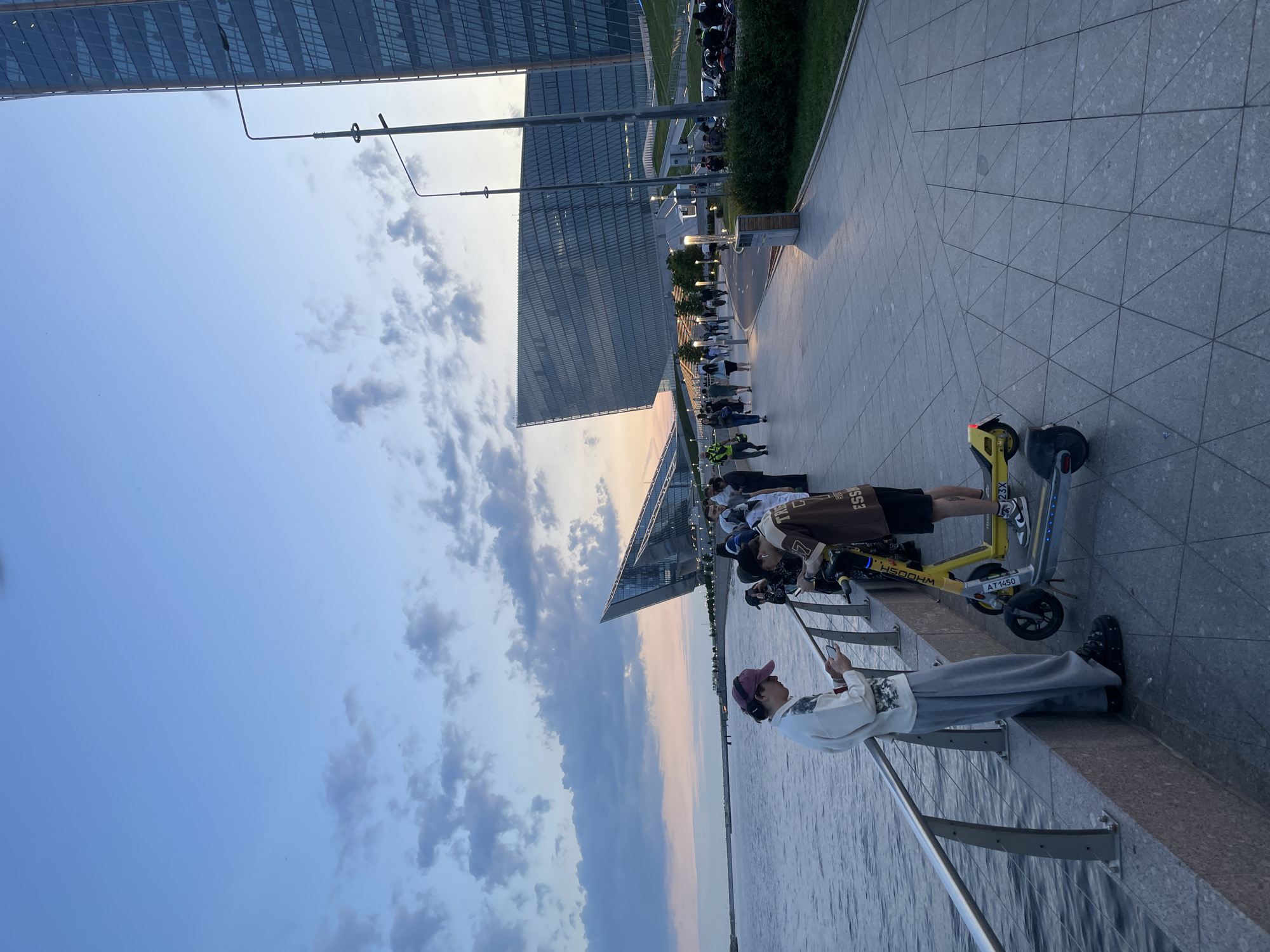 The image depicts a waterfront promenade in an urban setting at dusk. A body of water is on the left, bordered by a metal railing and a stone embankment where two individuals stand near electric scooters. The person on the left, wearing a maroon cap and a white sweatshirt, is looking at their phone. Beside them, another person in a brown and white patterned shirt is also looking at their phone. Two electric scooters, one yellow and one dark grey, are parked on the paved walkway.
Further along the promenade, a group of people are walking and standing. In the background, a modern architectural complex with distinctive angular glass buildings dominates the scene. The sky is a gradient of blue and soft orange hues, indicative of sunset, with scattered clouds. Streetlights and landscaping are visible along the promenade, and a tall glass skyscraper stands to the right. The overall impression is a contemporary urban scene during twilight hours.