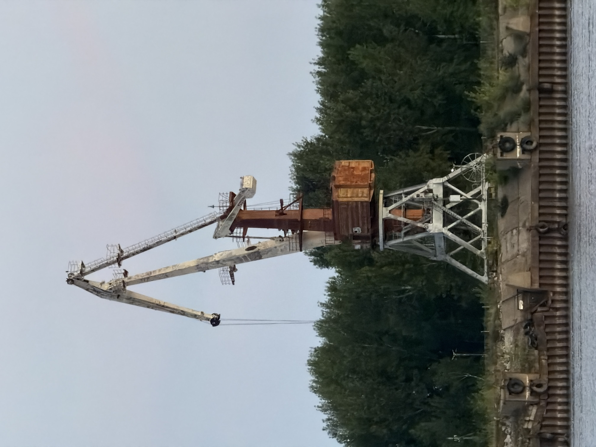 This image shows a large, industrial crane situated on the bank of a body of water, likely a river or canal. The crane is old and appears to be made of weathered metal, with rust visible on its structure. It has a tall, angled boom with a hook hanging down. Behind the crane is a dense line of green trees, suggesting a natural landscape bordering the industrial site. In the foreground, a concrete embankment with vertical wooden or metal pilings runs along the water's edge. The water itself is calm and appears grey or muted in color. The sky above is a pale, overcast blue. The scene conveys a sense of industrial decay and quietness. The specific location is identified as Chaikovsky, Russia. There are no people visible in the image, and the crane is stationary.