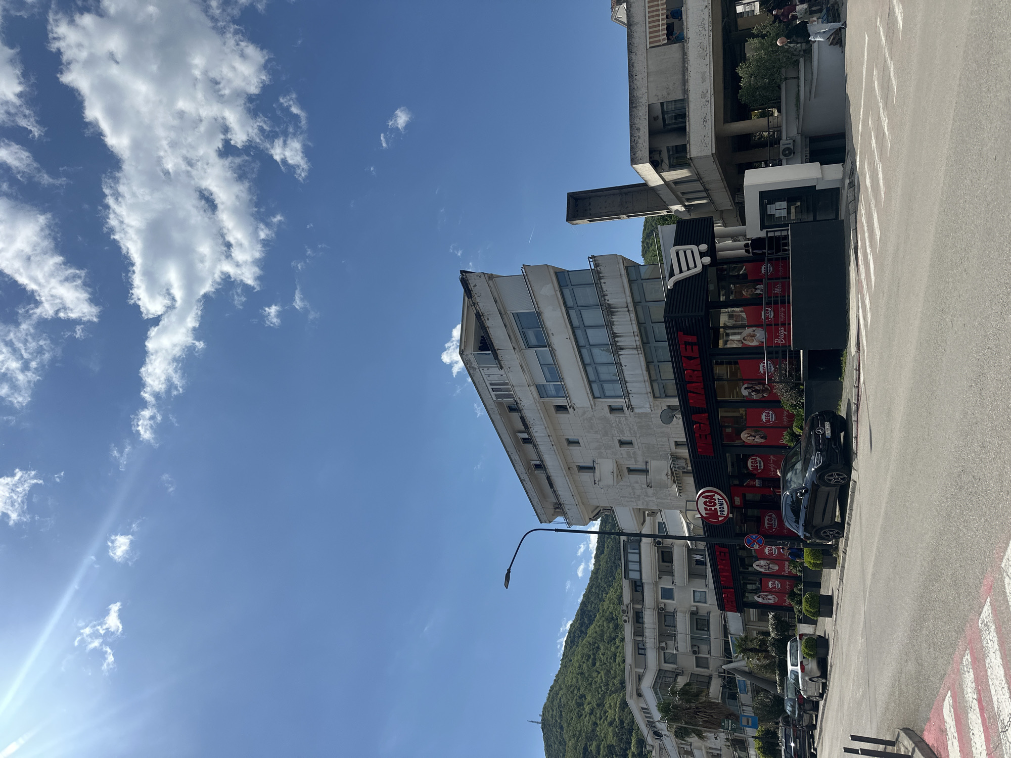 The image shows a street scene in Budva, Montenegro on a sunny day. The sky is blue with scattered white clouds. Prominently featured is a multi-story building with a "MEGA PROMET" supermarket on the ground floor. Cars are parked along the street adjacent to the supermarket. On the adjacent buildings, there are apartments with balconies. In the distance, a green hillside with trees is visible. The street is paved, and street markings are visible. There's a lamp post on the street.