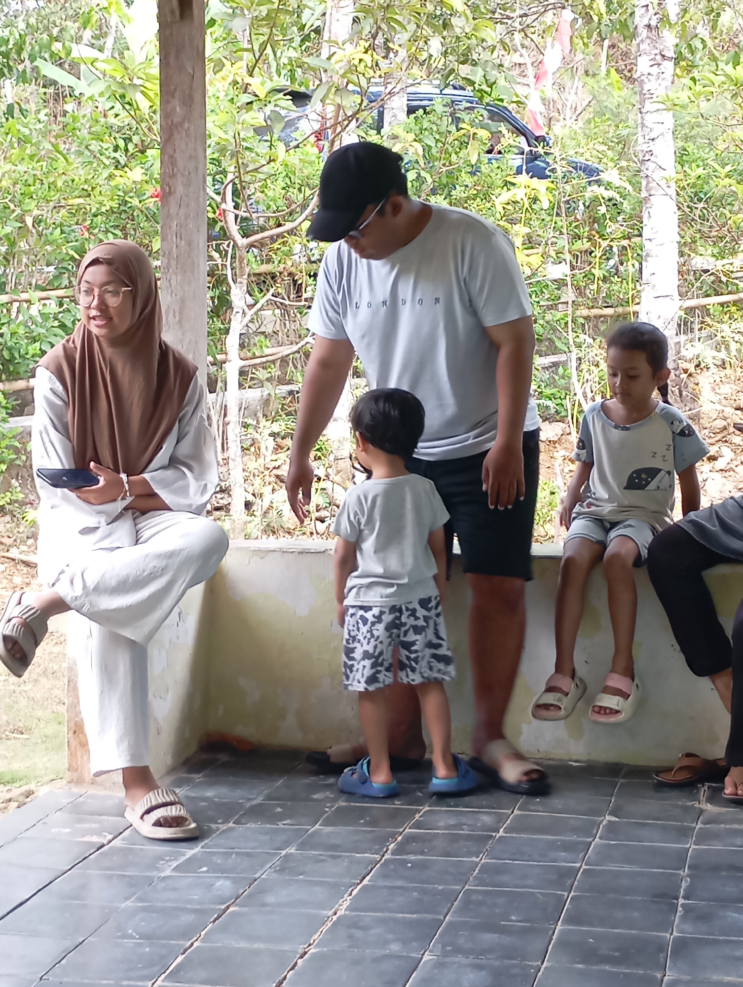 In Ketepung, Indonesia, a group of five people are gathered on a shaded tiled patio adjacent to lush greenery. A woman wearing a brown hijab and eyeglasses sits on a low wall, holding a dark mobile phone. To her right, a man in a grey t-shirt and black shorts stands, with a black baseball cap on his head. In front of him, a young boy in a grey t-shirt and patterned shorts stands, facing away from the camera. To the right of the man, a young girl in a grey t-shirt with a cartoon pattern and matching shorts sits on the wall. Partially visible on the far right is another person, likely an adult, wearing black pants and a grey shirt, with their legs bent. The background is dominated by dense foliage and trees, with a glimpse of a dark-colored vehicle in the distance. The overall impression is a casual, outdoor gathering.