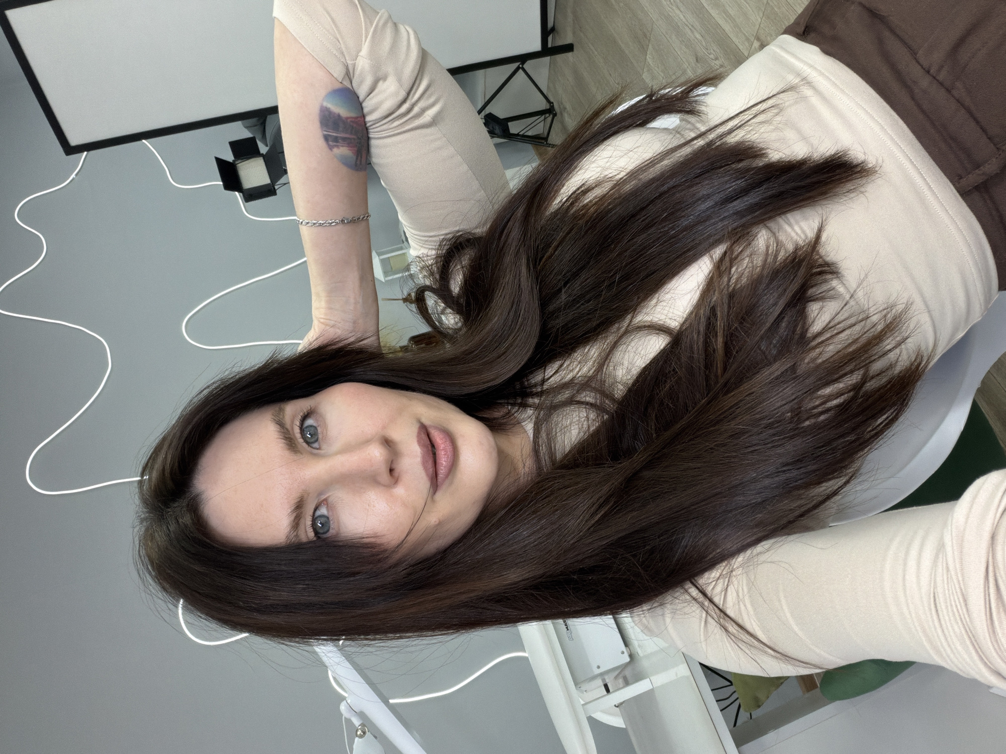 A Caucasian woman with long dark brown hair is pictured indoors. She has fair skin, blue eyes, and is wearing light-colored clothing. A tattoo of a landscape within a circle is visible on her left arm, along with a silver bracelet. The environment includes medical or dental equipment, suggesting a clinical setting. The background features photography lighting equipment. The image was taken in the city of Perm, Russia.