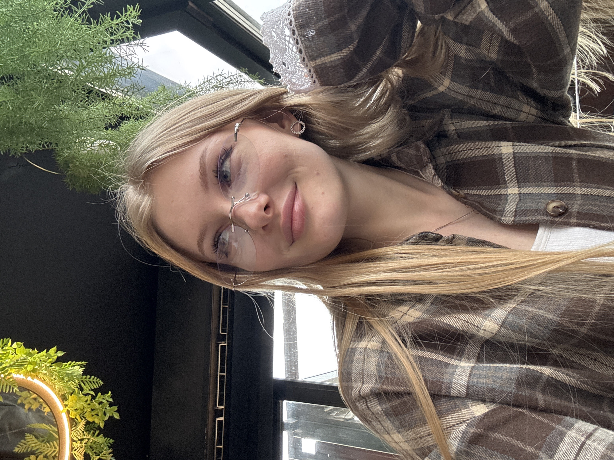 A photograph shows a young woman with blonde hair and eyeglasses in a brown plaid shirt. She is indoors, possibly near a window. There is greenery visible in the background, along with the window. The photograph may have been taken in a location in Perm, Russia.