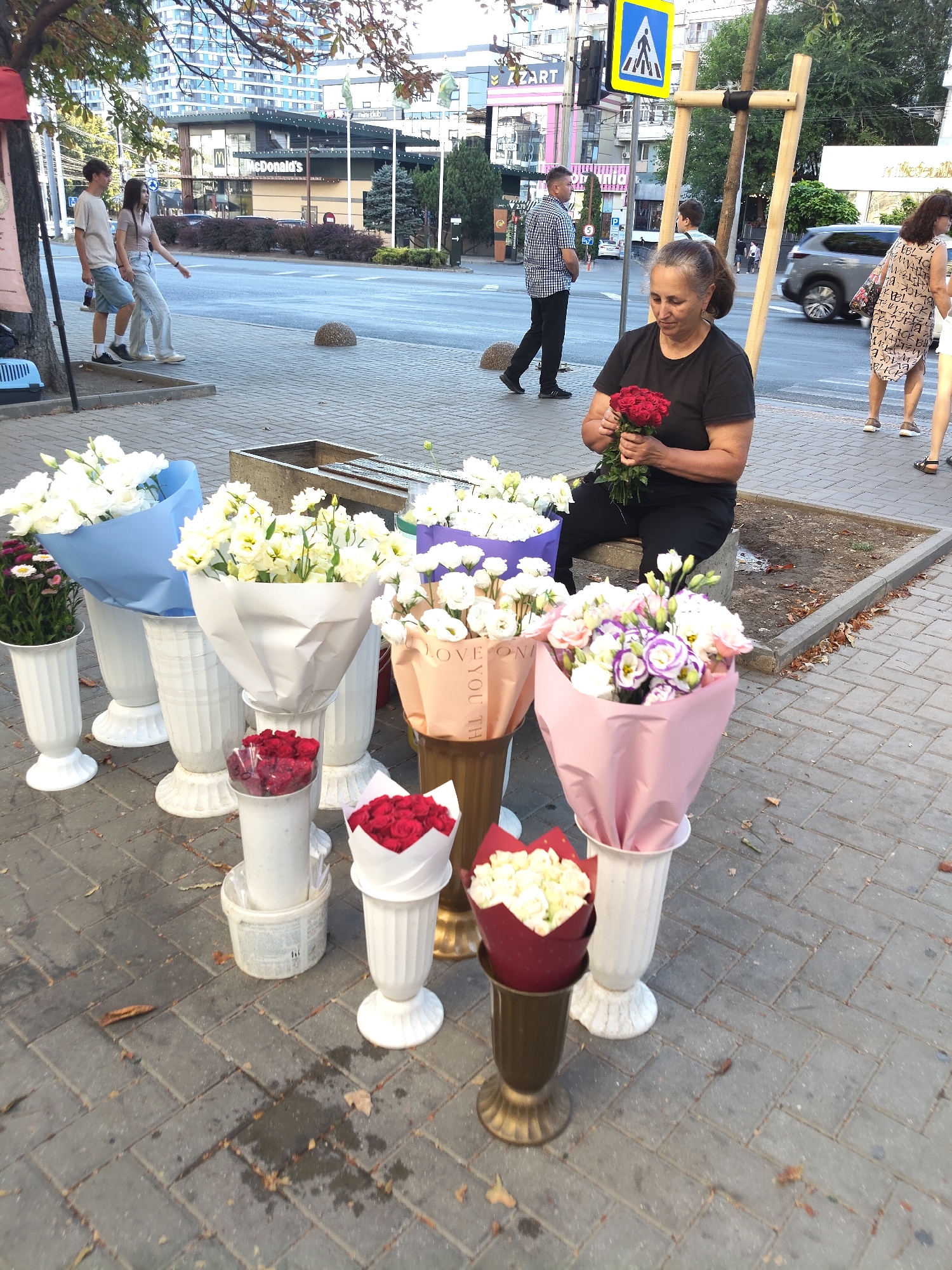 The image depicts a street scene in Chișinău, Moldova, featuring a woman selling flowers. The woman, seated on a concrete bench, holds a bouquet of red roses. Surrounding her are various arrangements of flowers in vases and containers, suggesting a flower stall. In the background, a McDonald's restaurant is visible, indicating an urban commercial area. Pedestrians are present in the scene, including a young couple walking by and another man standing on the sidewalk. The environment is paved with cobblestones and includes street furniture and foliage. The overall impression is that of a bustling city street with a flower vendor actively engaged in her trade.