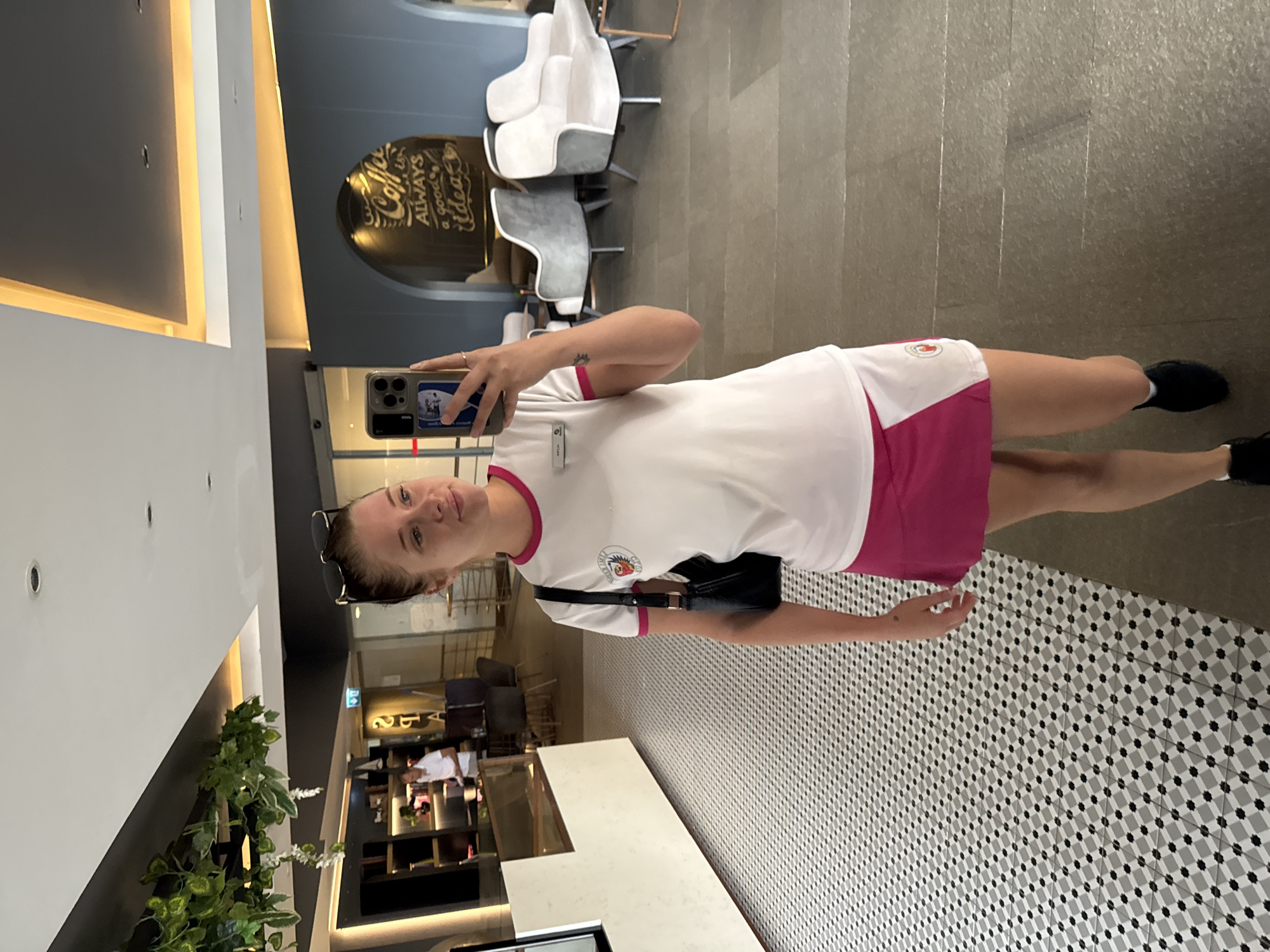 A young woman is taking a mirror selfie in what appears to be a hotel lobby or a similar establishment. She is wearing a white t-shirt with pink trim and a pink skirt. She has sunglasses on the top of her head and a black purse slung over her shoulder. The background shows seating areas, decorative elements, and what looks like a coffee-themed mural. The floor has a geometric patterned tile. The presence of a name tag on her shirt suggests she might be an employee of the establishment. The context of Aksu, Turkey, is not discernible from the visual information in the image.