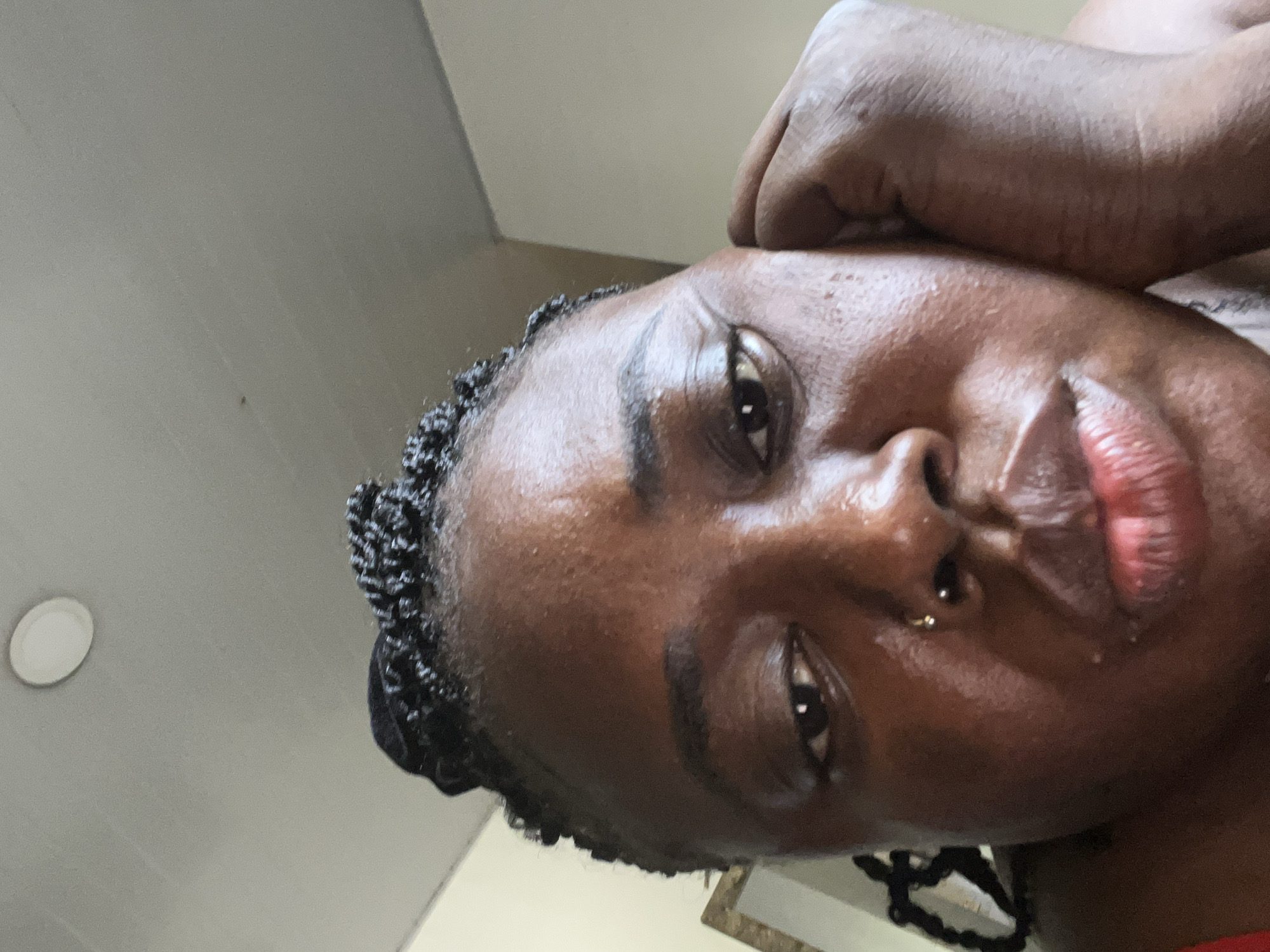 The media file shows a close-up, low-angle shot of a person's face, with their head tilted and a hand resting on their forehead. The person has dark skin and braided hair. They are wearing a gold hoop in their nose. Their lips are full and appear to be slightly moist, with a reddish tint. The background is a muted gray, with a round light fixture visible on the left side of the frame, and a hint of a wall and ceiling. The lighting suggests it is daytime, possibly indoors. The overall mood is intimate due to the close-up nature of the shot. There is no visible text in the image. The provided location context is Abuja, Nigeria.