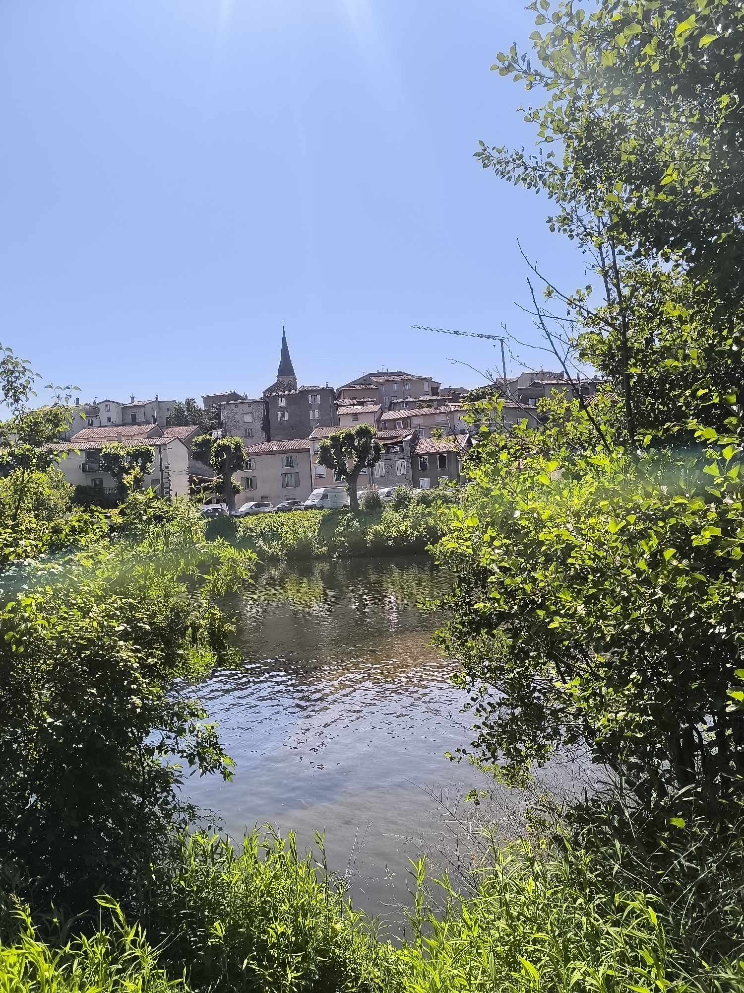 The image depicts a view of the town of Chézenas, France, framed by foliage on either side. A body of water, likely a river, occupies the foreground, reflecting the light and the sky. The town sits on a slight elevation, with buildings, including a church with a prominent steeple, visible against the clear blue sky. A construction crane is visible behind the houses on the horizon. Several cars are parked in front of the houses in the distance. The overall impression is of a sunny day in a picturesque, small French town.