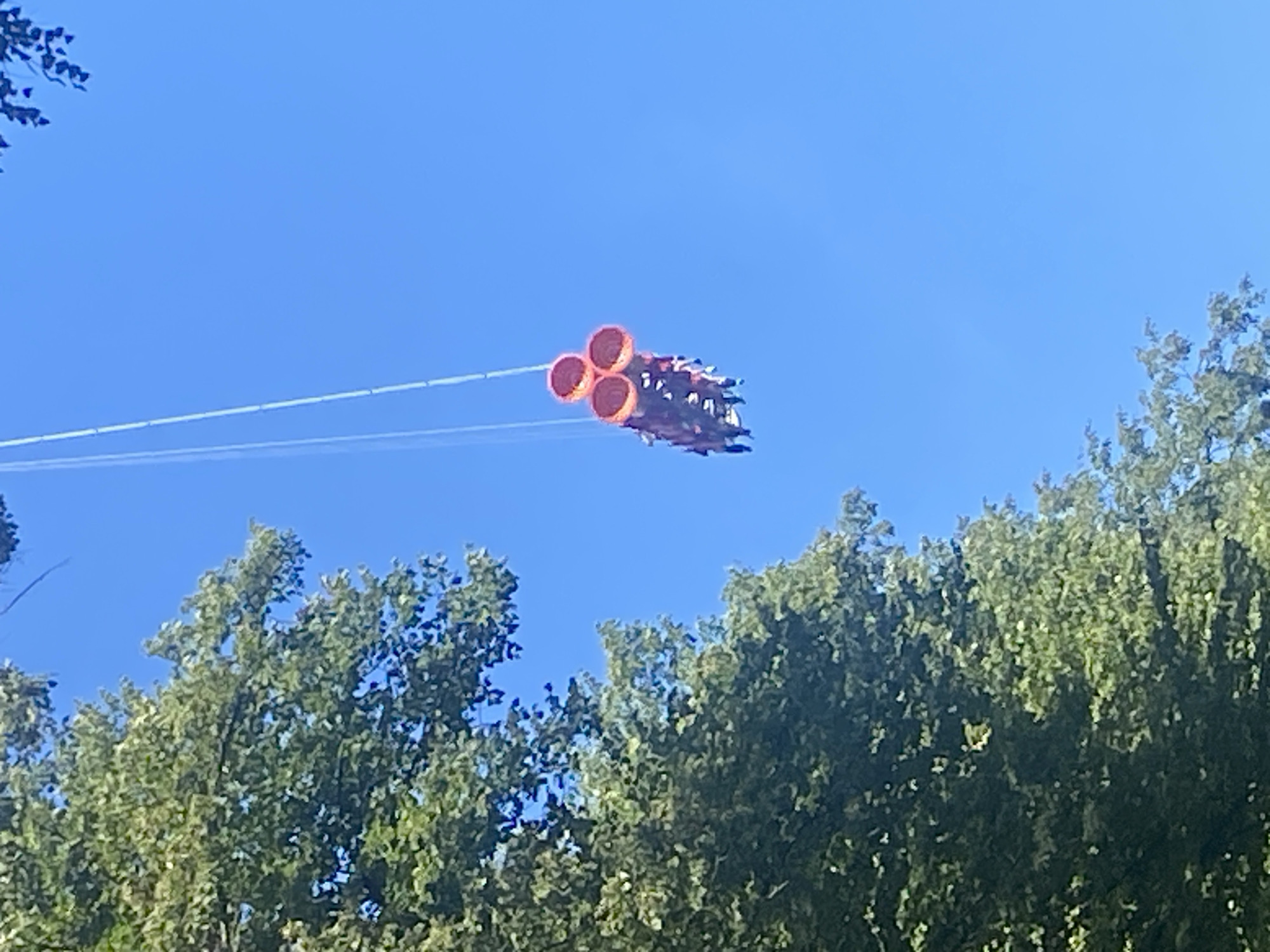 The image shows a thrill ride with multiple gondolas suspended in the air against a bright blue sky. The gondolas appear to be triangular in shape with three circular openings at the top, possibly representing seats or cabins. They are attached to a central support structure by cables. The ride is high in the air, with the tops of lush green trees visible in the lower left and upper right corners of the frame. The sunlight suggests it is a clear day. There are no visible landmarks that would indicate the specific location within Saint Petersburg, Russia. The scene captures a moment of excitement and adventure as people experience the thrill of the ride.