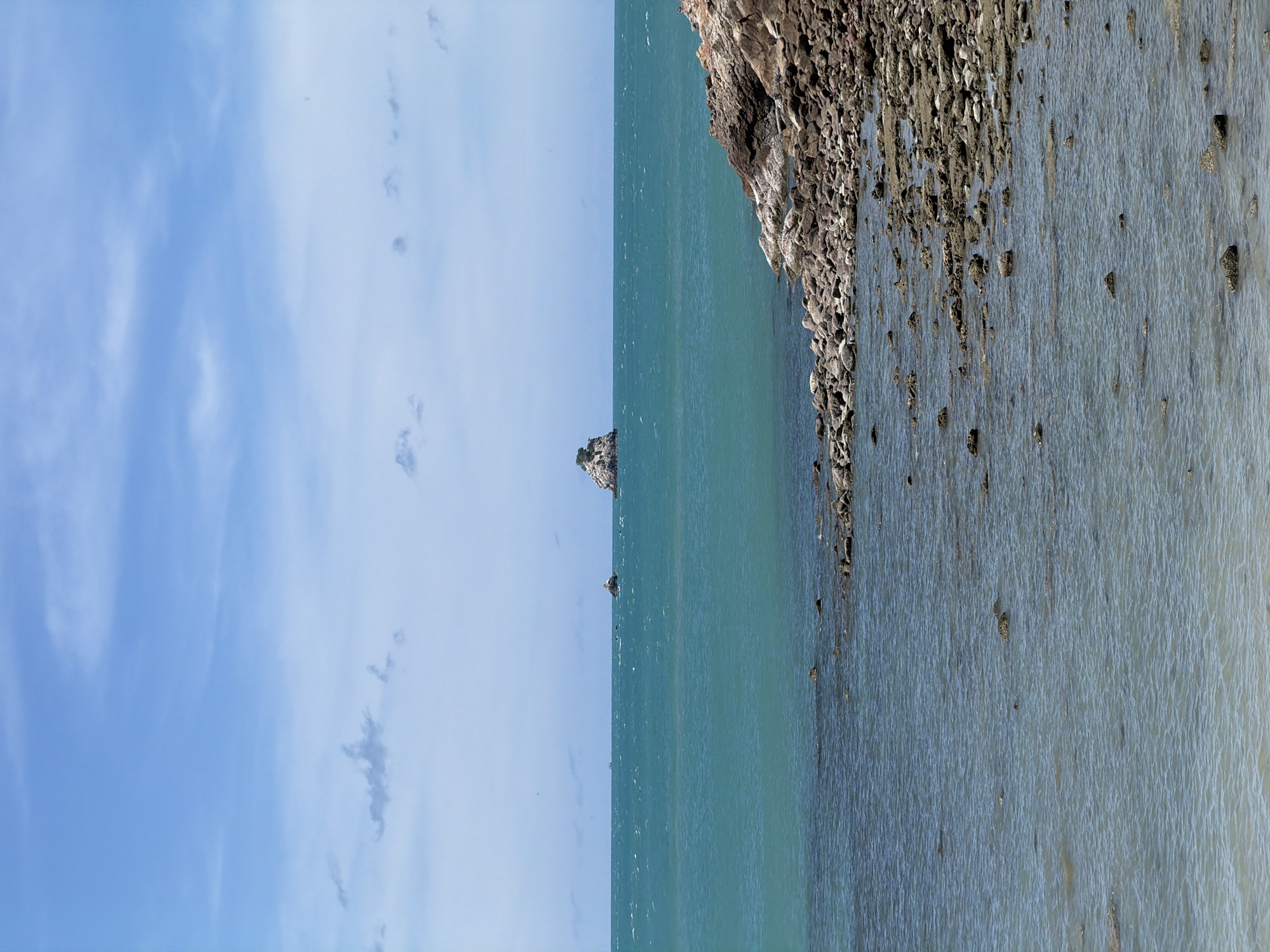 The image shows a panoramic view of the ocean with a rocky shoreline in the foreground. The water is a clear turquoise color and appears relatively calm, with small waves lapping at the shore. The shoreline is covered in various sizes of rocks and pebbles, with some larger boulders scattered throughout. In the distance, a small, tree-covered island is visible in the middle of the ocean. The sky is a light blue with scattered white clouds. The scene is likely located at Baan Plai Laem, Thailand, a coastal village known for its scenic beaches and rocky outcrops. There are no people or animals visible in the image.