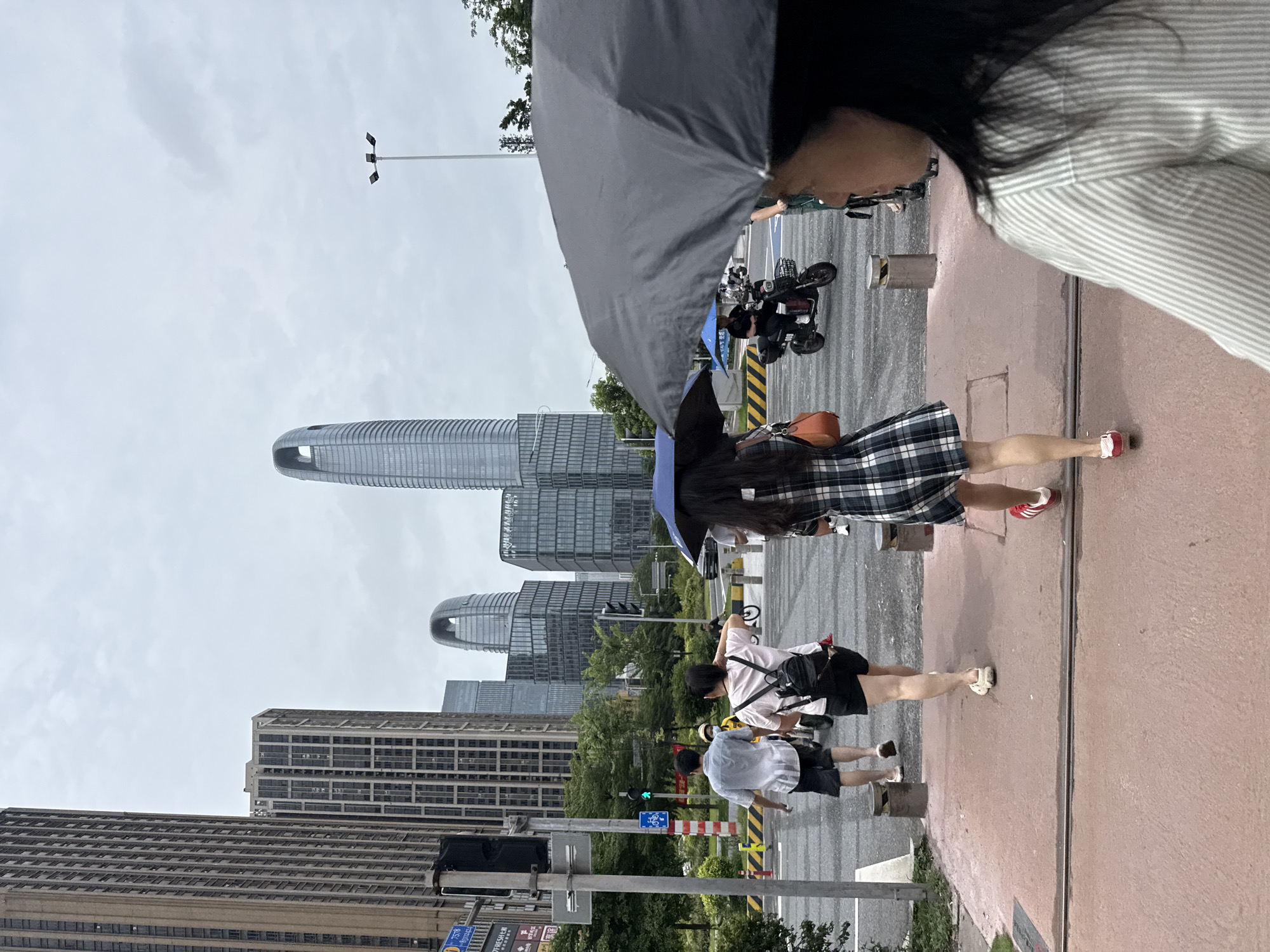 In Haizhu District, China, on a cloudy day, people are walking across a crosswalk in front of a modern cityscape. A man in a light-colored short-sleeved shirt and black shorts, and a woman in a pink t-shirt and black shorts with a backpack, are walking away from the camera towards the right. Another woman, also walking away from the camera and towards the right, wears a plaid dress and red shoes. A fourth person, whose face is visible in the foreground and who is holding a black umbrella, is walking towards the camera and to the right. In the middle ground, a person is riding a scooter and another is on a bicycle. Tall, modern buildings with glass facades and distinct architectural designs, including a curved-top skyscraper, dominate the background. Streetlights and traffic signals are visible. The crosswalk is marked with yellow and white stripes, and the ground is a reddish-brown color.