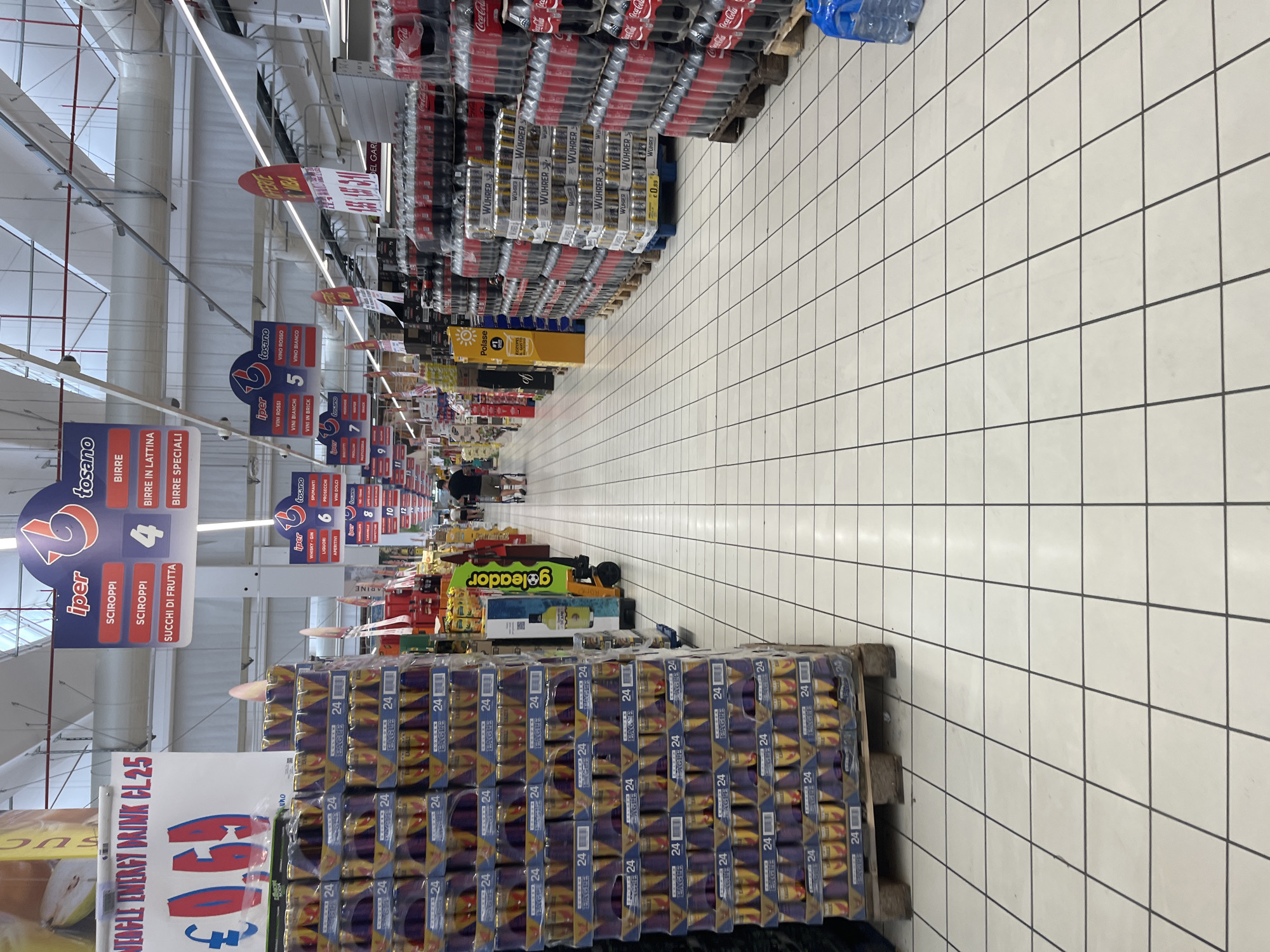 The image depicts the interior of a supermarket, likely an Iper Tosano store, given the signage. The perspective is from a low angle, looking down an aisle. On the left, stacks of canned beverages, possibly energy drinks, are prominently displayed on a pallet. These cans are labeled "24" and feature the "Eagle" branding. Further down the aisle, on the right, large stacks of bottled drinks, appearing to be Coca-Cola, are visible. Various promotional signs hang from the ceiling, advertising products and prices in Italian. One sign indicates "WEAGLE ENERGY DRINK CL 25" with a price of "€0.69". Another set of signs details product categories like "SCIROPP", "BIRRE", and "SUCCHI DI FRUTTA". People are present in the background, appearing to be shoppers, with one individual pushing a shopping cart. The floor is tiled with a white checkerboard pattern. The environment suggests a well-stocked retail space. Based on the typical locations of Iper Tosano stores, this scene could be within the city of Castiglione delle Stiviere, Italy, where an Iper Tosano supermarket is located.