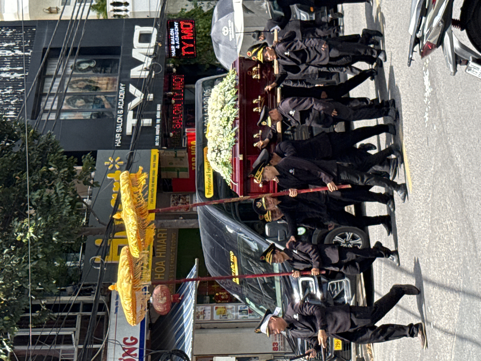 In Nha Trang, Vietnam, a funeral procession moves down a street lined with businesses. Six pallbearers, dressed in dark suits and hats with gold trim, carry a dark red coffin adorned with a large white floral arrangement. They are accompanied by two individuals holding decorative yellow parasols on tall poles. Behind them, a black van with tinted windows is visible. The businesses in the background include "TY MO HAIR SALON & ACADEMY" and "HOA HMART." The street is paved and appears to be a sunny day with shadows cast on the ground.