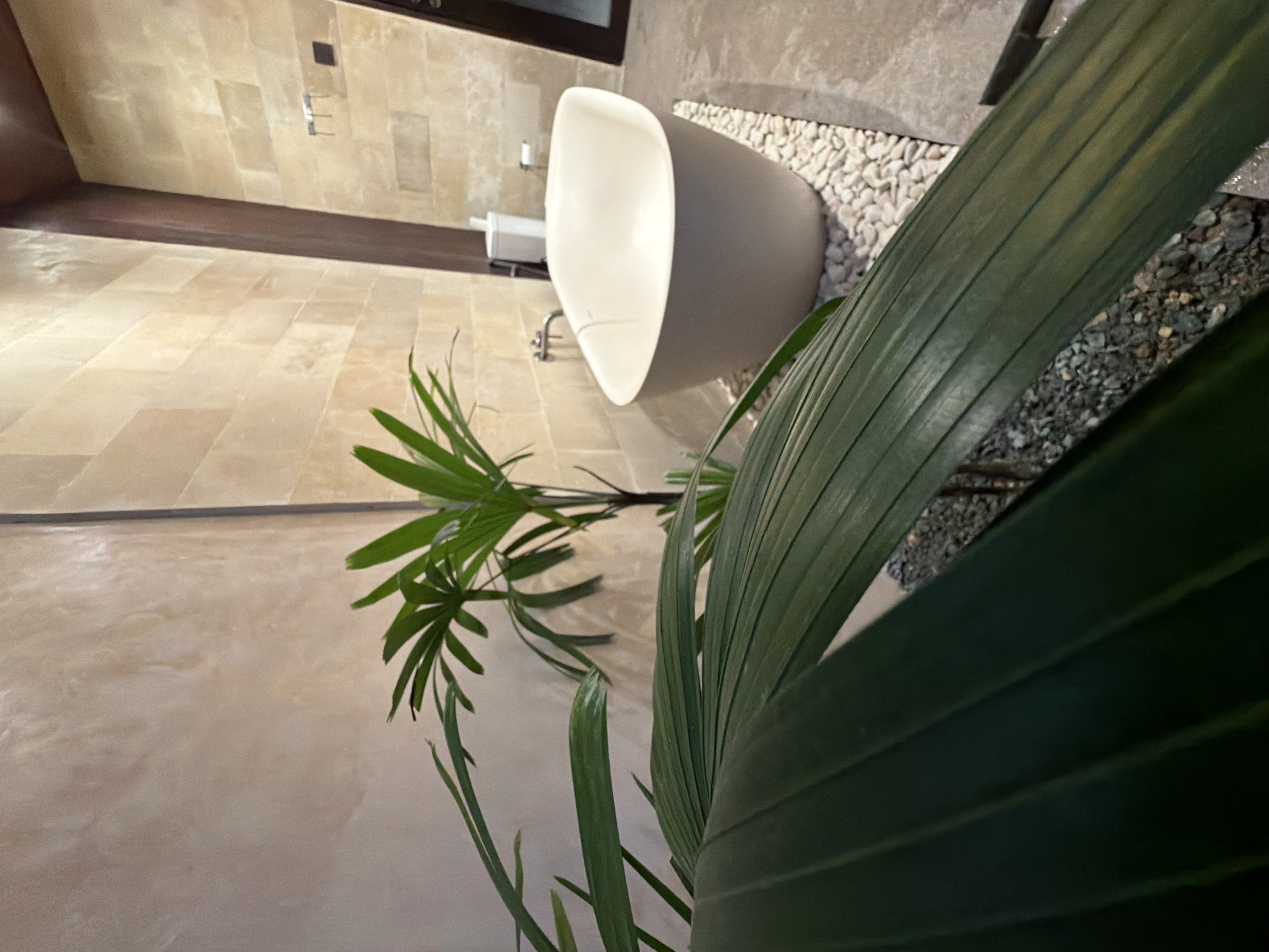 A free-standing, egg-shaped bathtub with a light gray exterior and white interior is situated in a bathroom with light beige tiled walls. The tub rests on a bed of small, white pebbles. To the left of the tub, a tall, thin object, possibly a white trash can, and a roll of toilet paper are visible. A silver faucet is mounted on the wall behind the tub. In the foreground, large, dark green palm leaves obscure a significant portion of the view. The leaves are positioned in front of a potted palm plant. The floor appears to be a mix of concrete and gravel. The overall ambiance suggests a modern, spa-like bathroom. This scene is located in Kuta, Indonesia.