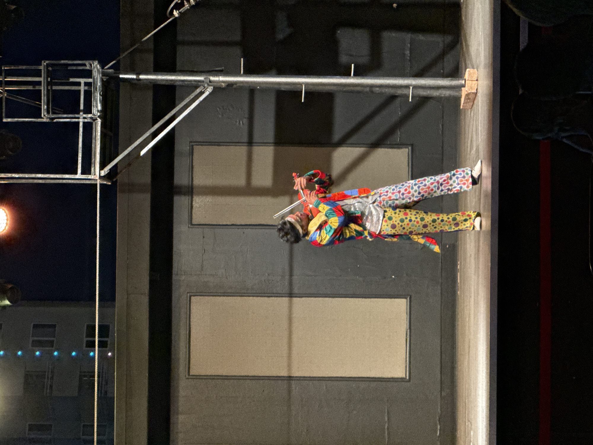 A person dressed as a clown stands on a stage, playing a violin. The clown is wearing a colorful, patchwork-style jacket with a red, yellow, blue, and green color scheme, and brightly patterned pants with yellow, pink, and blue circles. The clown also has a white headband. The stage has a dark gray backdrop with two large, light brown rectangular panels. To the right of the stage, a metal scaffolding structure rises, with a bright orange light fixture visible at the top. A brick rests on the stage floor near the base of the scaffolding. In the background, a building with multiple windows is illuminated by blue lights. The scene appears to be an outdoor performance space at night, given the lighting and dark sky.