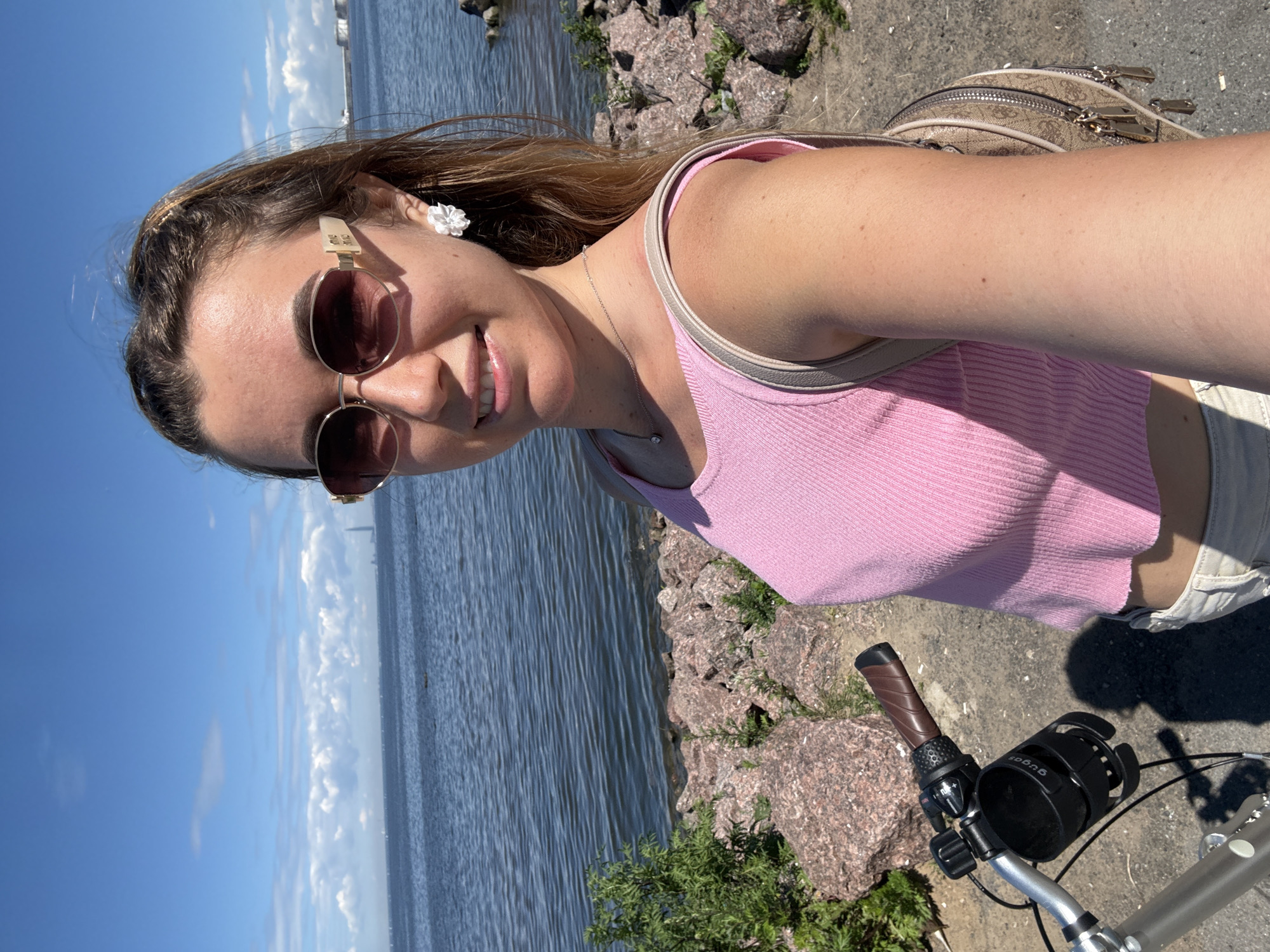 A young woman with brown hair pulled into a ponytail and wearing sunglasses, a pink top, and light-colored shorts stands on a paved path next to a body of water. She is holding a bicycle with a black cup holder attached to the handlebars. The background shows a calm blue body of water with a few sailboats and a distant cityscape. The environment is sunny with scattered white clouds in a bright blue sky. The path is lined with large rocks and some green vegetation.
