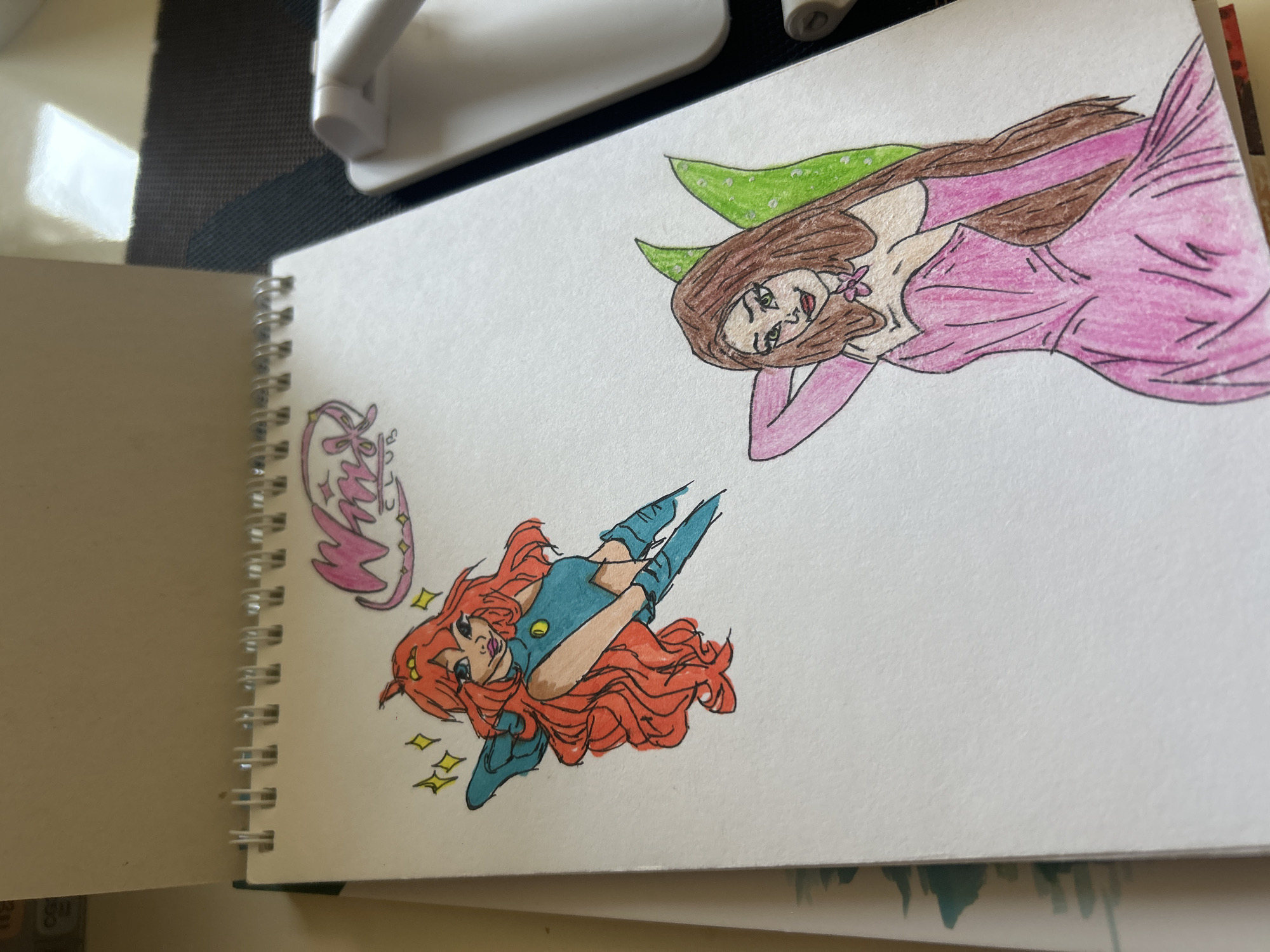 The image shows a close-up of a sketchbook open to a page with two hand-drawn and colored illustrations of cartoon fairies. The illustration on the left depicts a fairy with long, wavy orange hair, blue eyes, and a blue outfit. She has yellow sparkles around her head and is posed in a dynamic, flying stance. Above her head is a pink and purple logo with the words "WINX CLUB". The illustration on the right shows a fairy with long brown hair, green eyes, and a pink dress with long gloves. She has green fairy wings and is posed with one arm behind her head. The sketchbook is bound with silver spiral wire and rests on a dark, textured surface. A white object, possibly a desk lamp or phone stand, is visible in the background to the right, partially out of focus. The environment appears to be an indoor setting, likely a desk or table. The image does not provide any information or context related to the city of Khyrdalan, Azerbaijan.