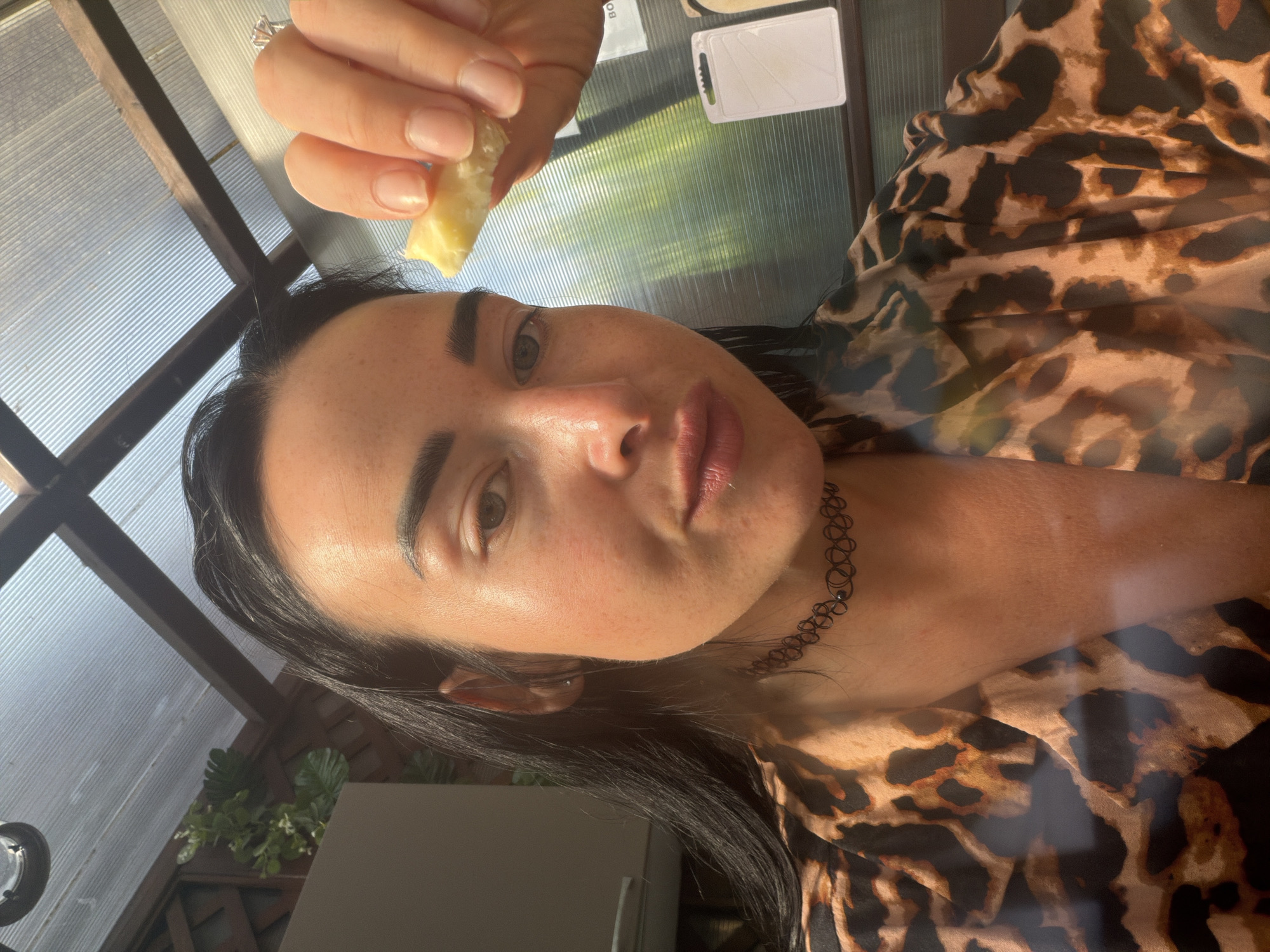 A woman is taking a selfie, holding a piece of cheese near her face. She is wearing a leopard-print shirt and a black choker necklace. Her right hand is in focus, showing a diamond ring on her index finger. The background appears to be an outdoor or semi-outdoor setting, possibly a covered patio or greenhouse, with a corrugated plastic roof. In the distance, there is greenery visible through the translucent roof. The overall lighting suggests it is daytime. The location is specified as Sochi, Russia.