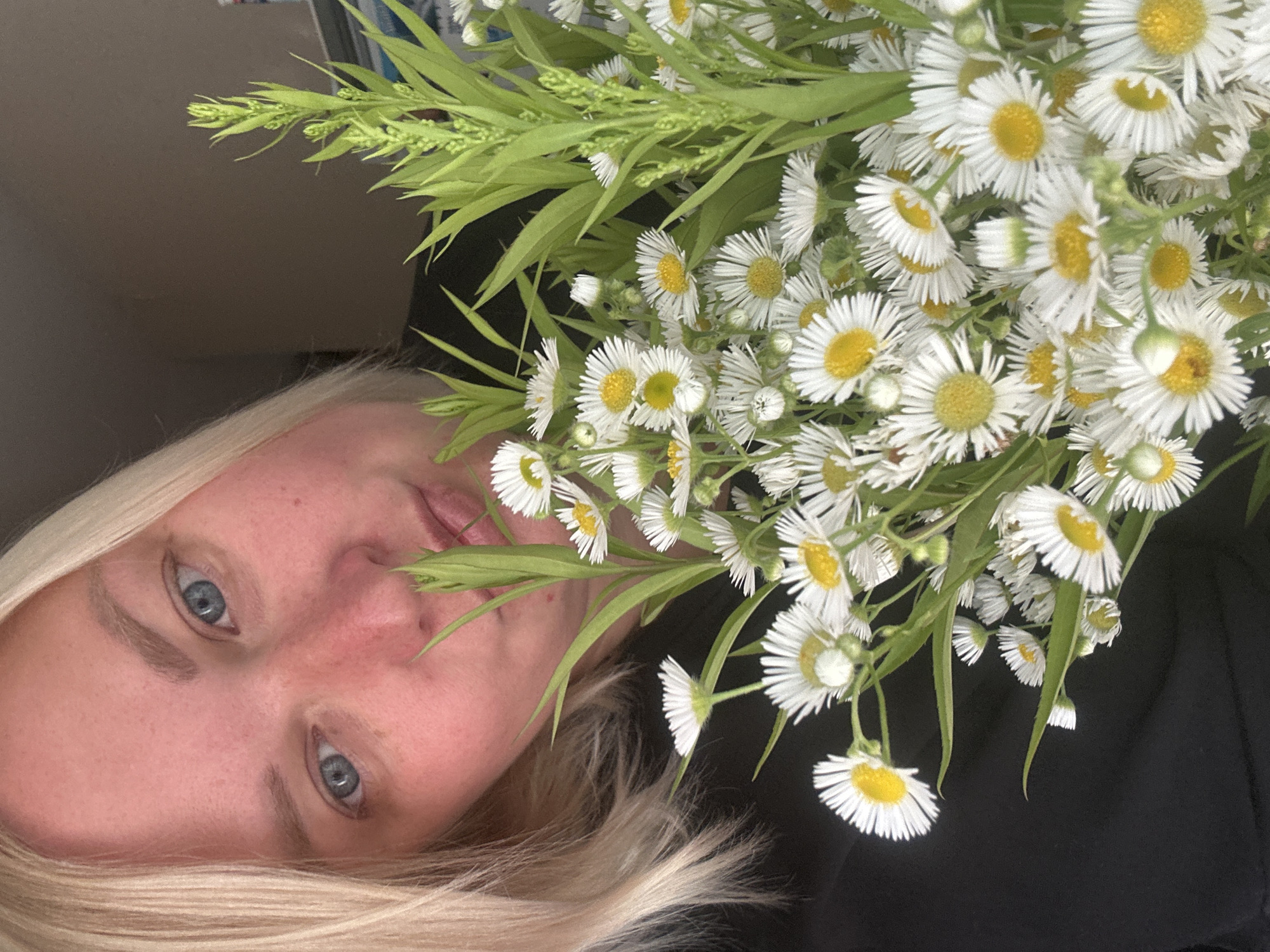 A close-up shot shows a blonde woman with blue eyes holding a bouquet of small white daisies with yellow centers. Her head is tilted, and she is looking towards the camera, with her lips slightly parted. The flowers are in focus and take up a significant portion of the frame, obscuring part of her face and chest. The background is dark and indistinct, suggesting an indoor setting. The image does not provide any information about the specific location within Chaikovsky, Russia.